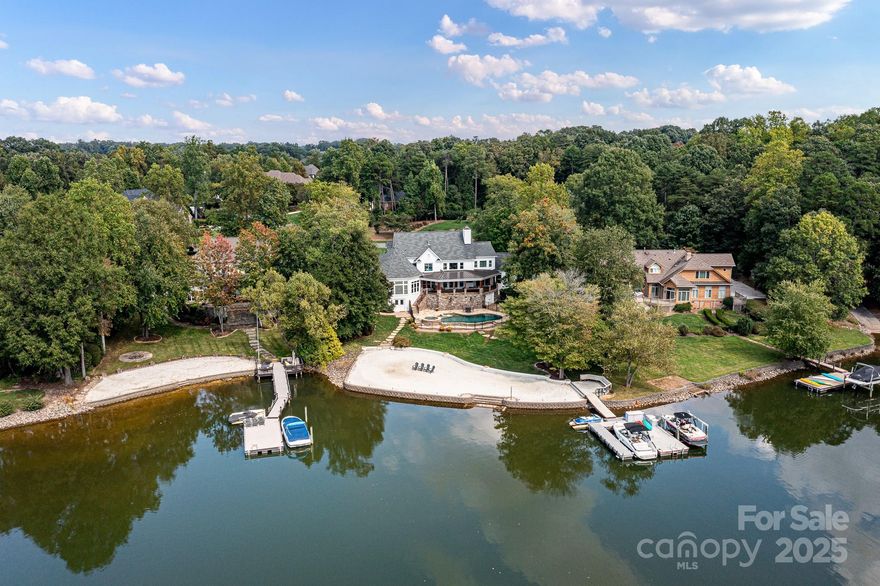 Located in the prestigious Patricks Purchase neighborhood on Lake Norman, this luxurious waterfront home embodies elegance and tranquility. Its stately white brick exterior, accented by natural wood shutters and lush landscaping, sets the tone for exceptional lakeside living. Inside, an open floor plan highlights vaulted beam ceilings, a chef’s kitchen with custom cabinetry, and a light-filled great room with stunning water views.
Outdoor spaces are unparalleled, featuring a spacious covered deck with stone flooring, a resort-style pool and spa with pool house, and a private dock equipped with boat slips and jet ski ports. A sandy shoreline provides the perfect spot for lakeside relaxation. Nestled on a sprawling lot with mature trees and direct main channel access, this home offers the ideal combination of luxury, privacy, and recreational opportunities. Whether entertaining or enjoying peaceful moments by the lake, this property captures the essence of upscale lakeside living.