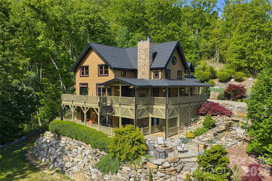 A one-of-a-kind private mountain estate, Bearadise Mountain Lodge sits on 14.69 wooded acres with breathtaking long-range views of the Blue Ridge Mountains. Crafted with post & beam log construction and exceptional attention to detail, this 3-story, 5,000+ sq ft retreat offers an extraordinary blend of rustic elegance and relaxed luxury.
The gentle sounds of the custom native stone waterfall and pond invite you down stone steps—accented by custom railings designed by a local artist—to scenic overlooks and thoughtfully curated outdoor living spaces. From the stone patio and fire pit to the multi-level covered and sun-drenched decks, every setting is designed to capture sweeping mountain views and unforgettable sunsets. Inside, the main living area welcomes you with open-beam, tongue-and-groove vaulted ceilings, rich cherry hardwood floors, and a dramatic two-story wood-burning stone fireplace. The well-appointed kitchen with a generous island and eating bar flows seamlessly into the dining area, perfectly positioned to take in spectacular mountain vistas. The main-level primary suite offers a private wooded retreat, complete with a wall-to-wall custom built-in, dual walk-in closets, and a spa-inspired bath featuring a glass-enclosed dual-head shower and jetted soaking tub. Upstairs, an open loft with theater-style seating leads to two bedrooms, two full baths, a spacious family/game room, and two additional flexible-use rooms. The fully finished, light-filled lower level offers a private apartment with its own entrance—featuring a full kitchen, living and dining area with propane fireplace, spacious bedroom, luxury bath, and separate laundry—ideal for guests, multi-generational living, or rental potential. French doors open to a covered patio with outdoor dining and a classic porch swing. A freestanding 1,506 sq ft insulated, heated and cooled workshop provides exceptional versatility for hobbies, studio space and storage with easy entry vehicle access and ample parking. Located just 2 minutes from downtown Weaverville and 17 minutes from Asheville, this exceptional mountain property offers a rare combination of privacy, craftsmanship, and convenience. Don’t miss this rare opportunity to own a true mountain jewel!