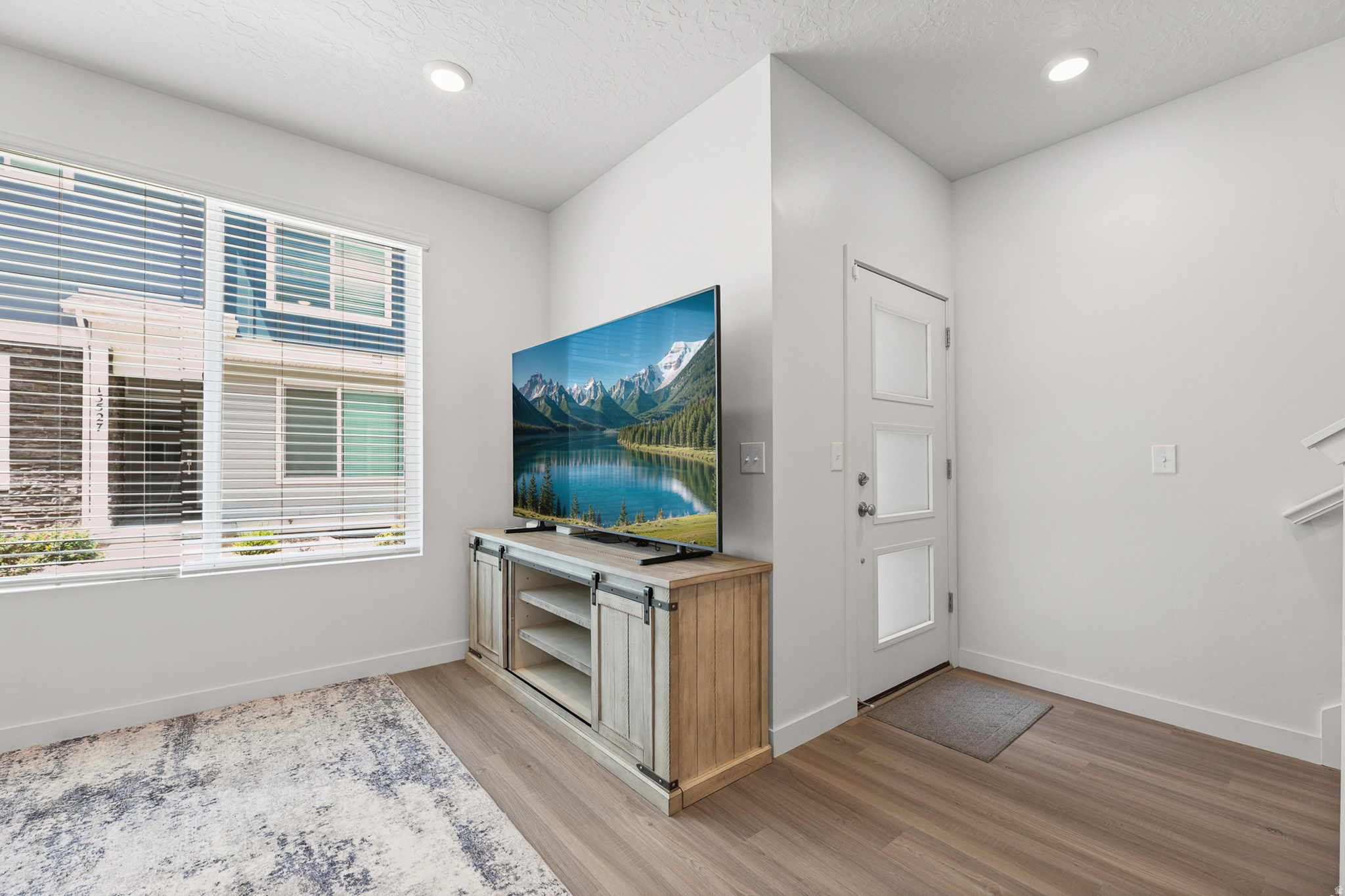 Living area featuring wood-finish flooring, recessed lighting, a large window with blinds, and a white entry door with frosted glass panels