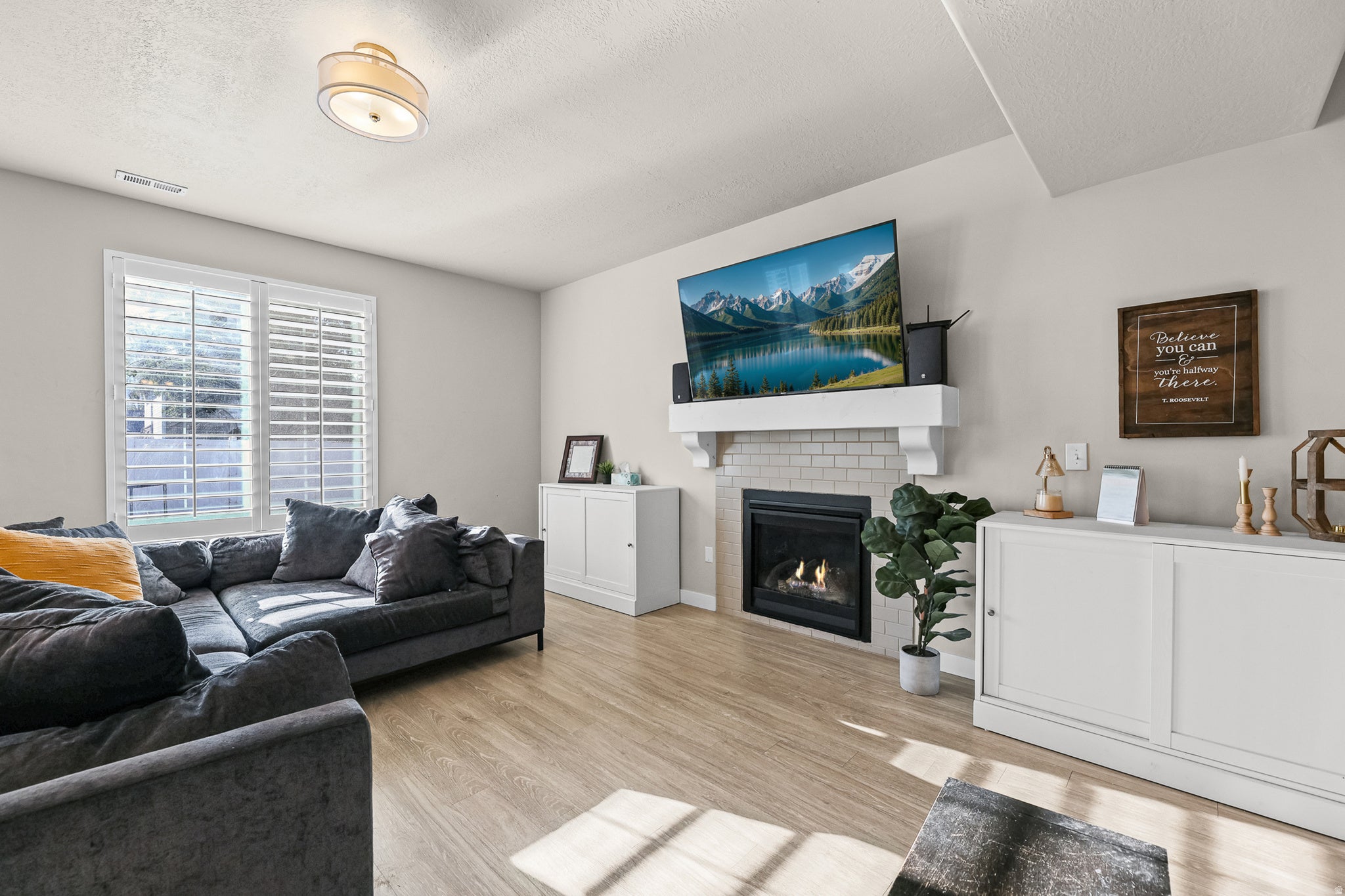 Living area featuring wood-finish flooring, a white mantel above a fireplace with white subway tile surround, and a large window with white plantation shutters