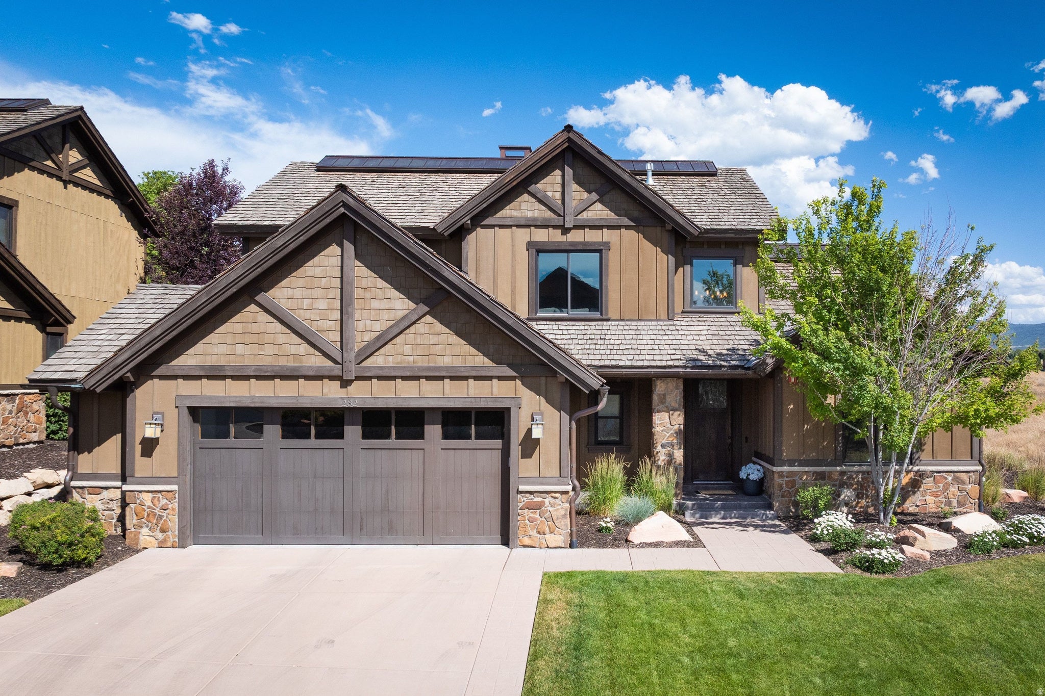 Craftsman-style home featuring a two-car garage, stone and shingle siding, a paved driveway, and a landscaped front yard with mature trees