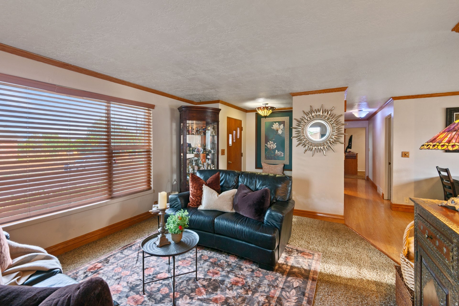 Bright living room showcasing hardwood flooring, a dark leather sofa, decorative sun-wall clock, and large horizontal-slat windows allowing abundant natural light