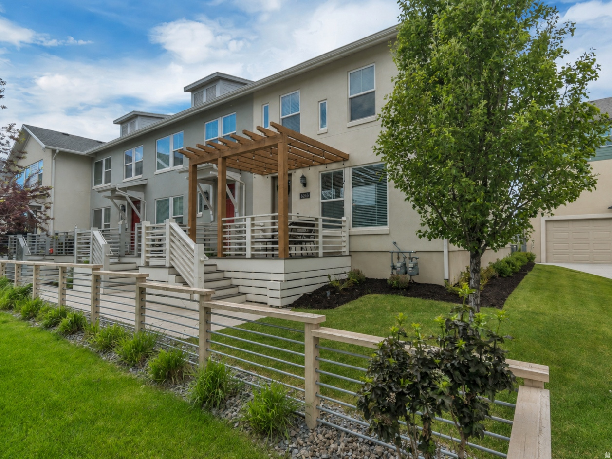Contemporary townhome exterior featuring stucco siding, multiple windows, a private deck with a wooden pergola, a two-car garage, and a manicured lawn