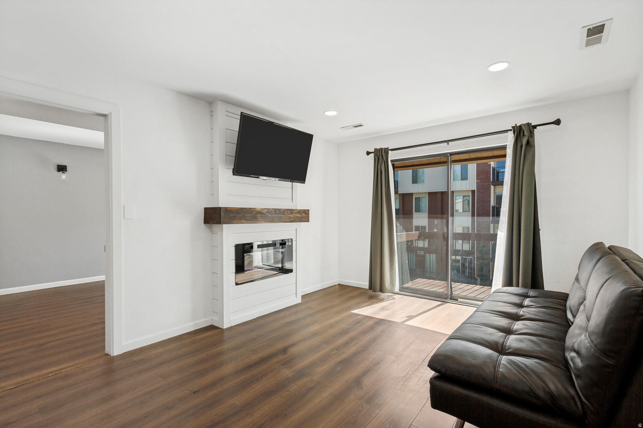 Living area featuring wood-finish flooring, a contemporary fireplace with a shiplap surround and wood mantel, recessed lighting, and a glass sliding door leading to a private balcony