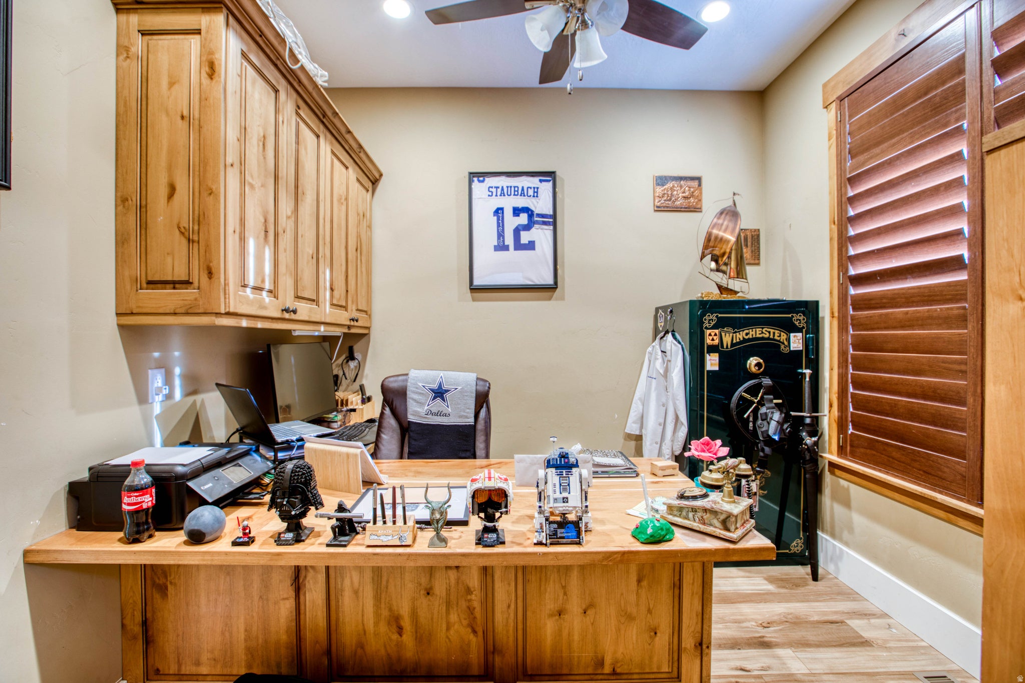 Built-in wood-finish cabinetry, wood-finish desk, wood-finish flooring, ceiling fan with integrated lighting, and wood-finish louvered shutters