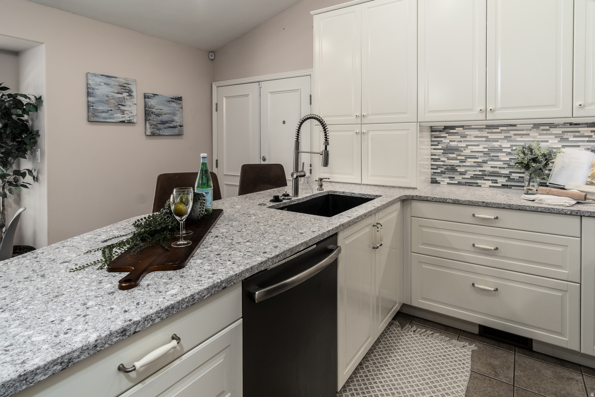 Kitchen featuring white shaker-style cabinetry, a dark undermount sink with a gooseneck faucet, light speckled countertops, a mosaic tile backsplash, and a built-in dishwasher