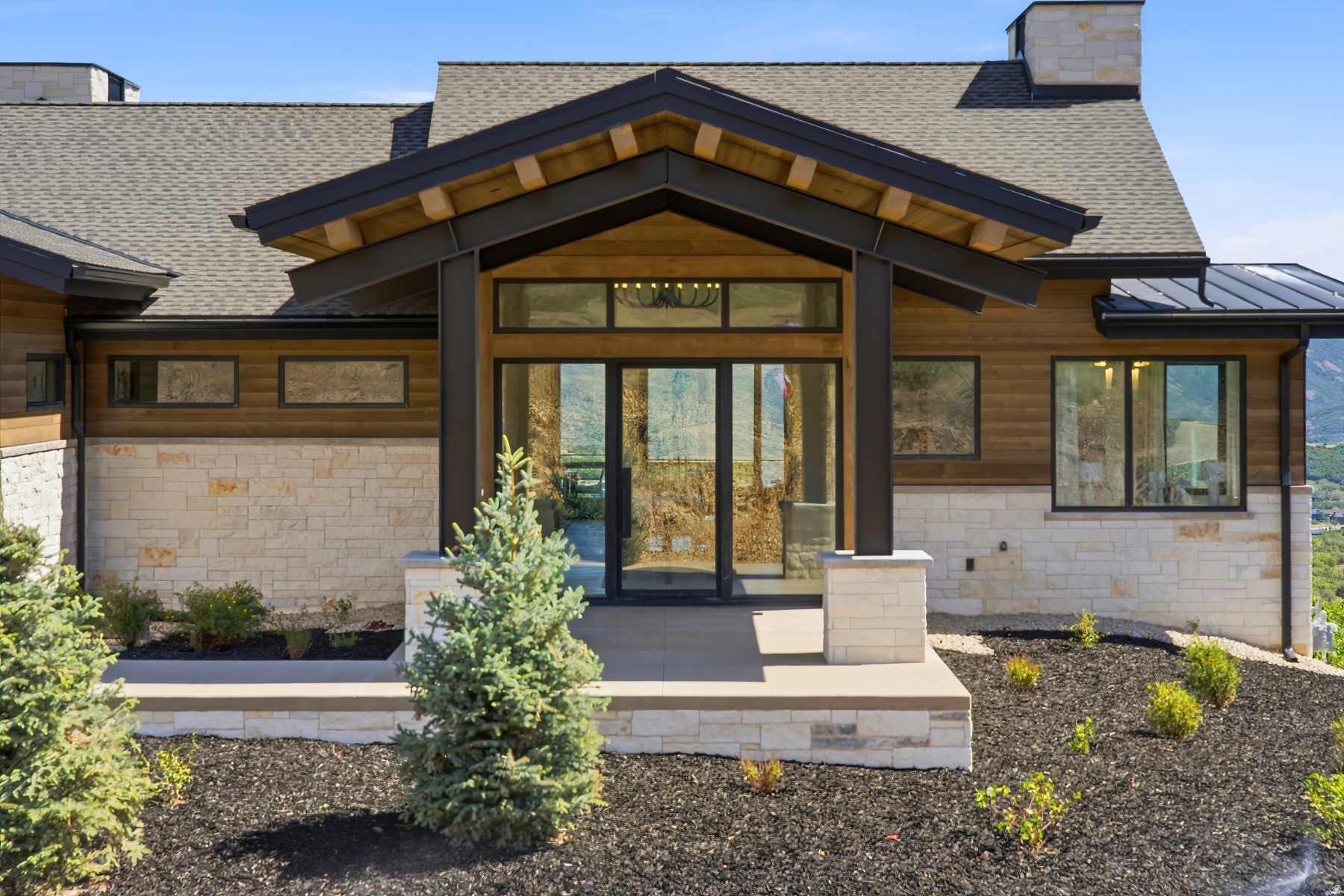 Contemporary facade featuring a stone and wood-finish exterior, covered entry with exposed beams, glass double-door entry, and a paved walkway