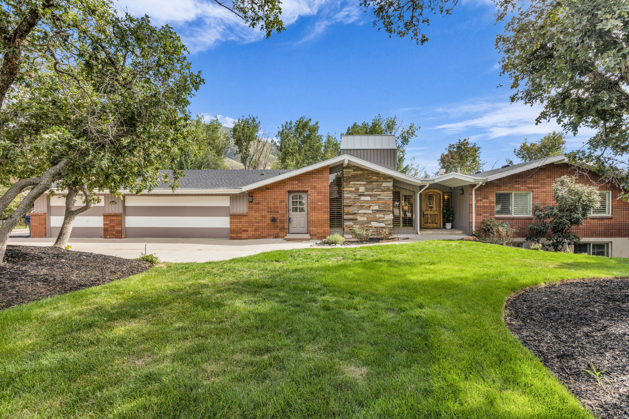Brick and stone facade, multi-car garage with striped doors, wood-finish entry door, and a landscaped front yard with mature trees