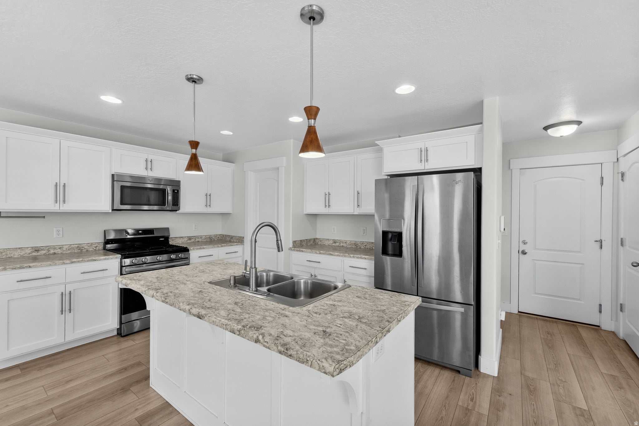 Attractive kitchen showcasing clean white cabinetry and quality stainless steel appliances for a contemporary aesthetic. The centerpiece kitchen island with dual-basin sink enhances both functionality and style, while beautiful wood-finish flooring adds w