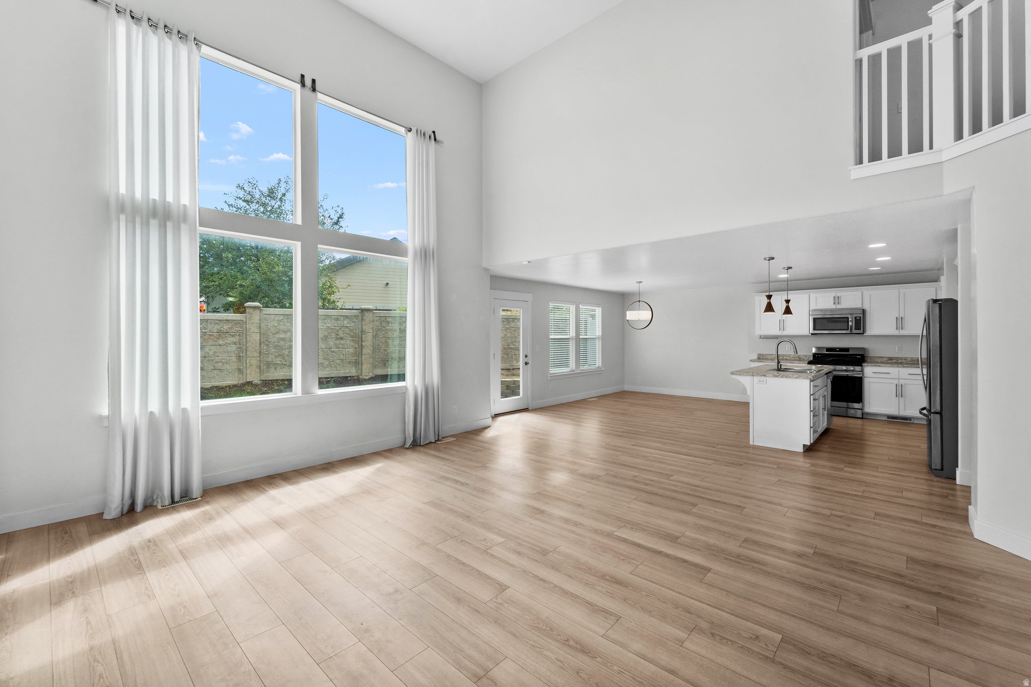 Stunning two-story living space showcasing dramatic high ceilings and abundant natural light from expansive windows. Warm wood-finish flooring complements the open layout, while the thoughtfully designed kitchen island with pendant lighting and quality st