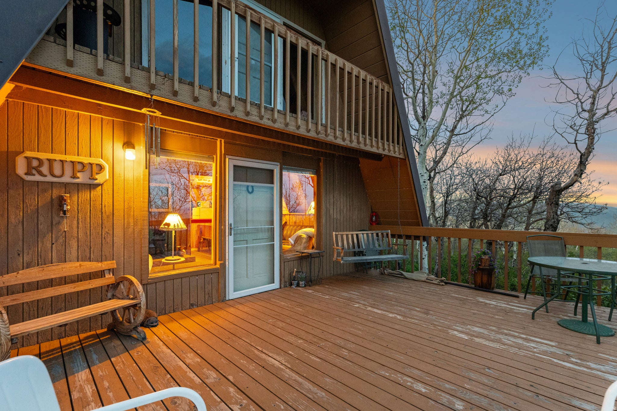 Front Deck at Dusk - Covered entry beneath the A-frame with a sliding glass door, porch swing, and bench seating, with interior lighting visible through oversized windows.
