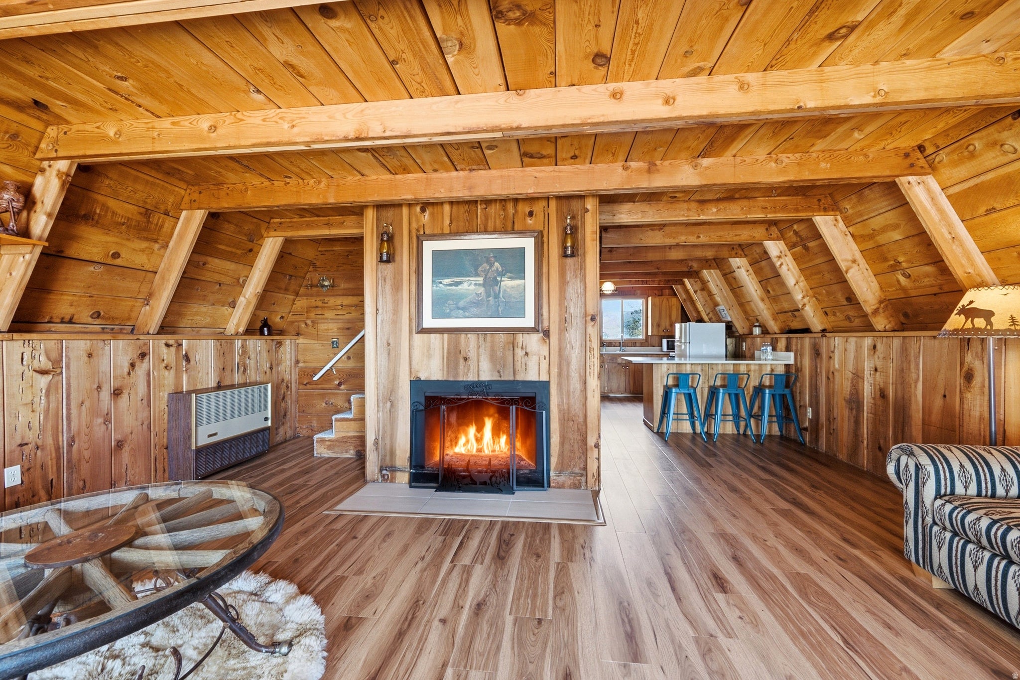 Great Room - Open living space framed by exposed wood beams and tongue-and-groove ceilings, with a wood-burning fireplace set into a full-height wood surround. Plank flooring carries through to the kitchen and dining area.