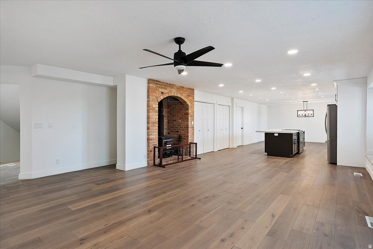Spacious open-concept room featuring wide plank wood-finish flooring, a brick archway with an interior fireplace, a kitchen island with a light-toned countertop, and recessed ceiling lighting