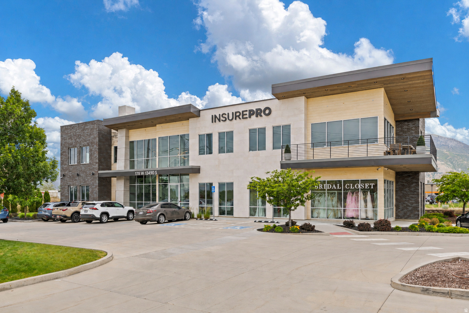 Contemporary commercial building featuring a stone and stucco facade, large plate glass windows, a second-story balcony with metal railings, and a prominent mountain backdrop