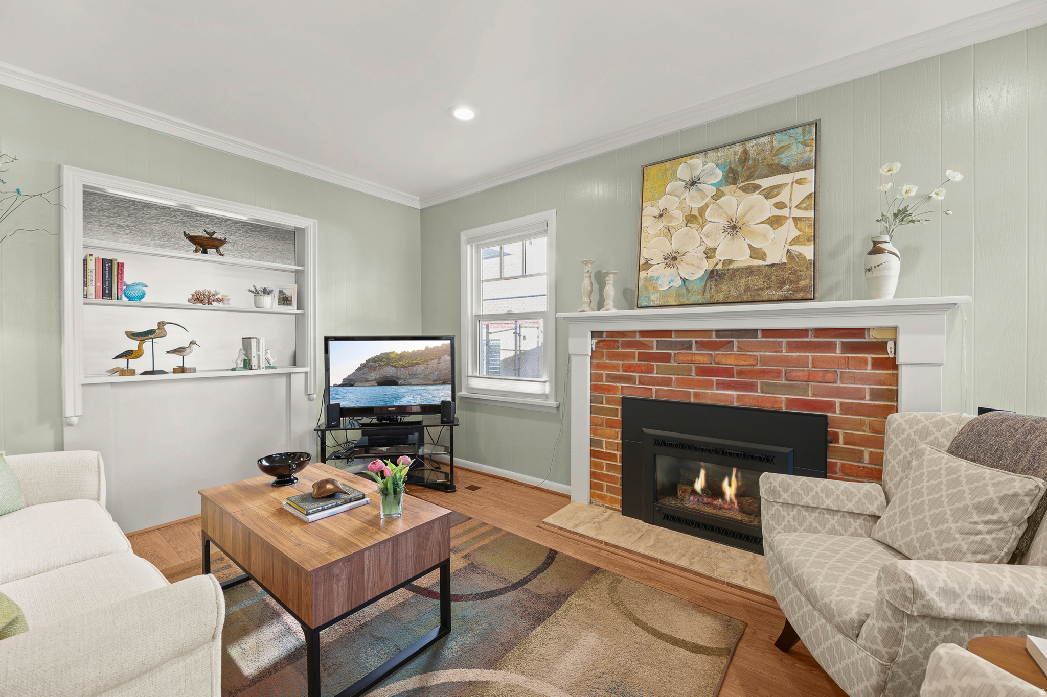 Living space featuring a brick-surround fireplace with a white mantel, built-in shelving, hardwood flooring, and crown molding