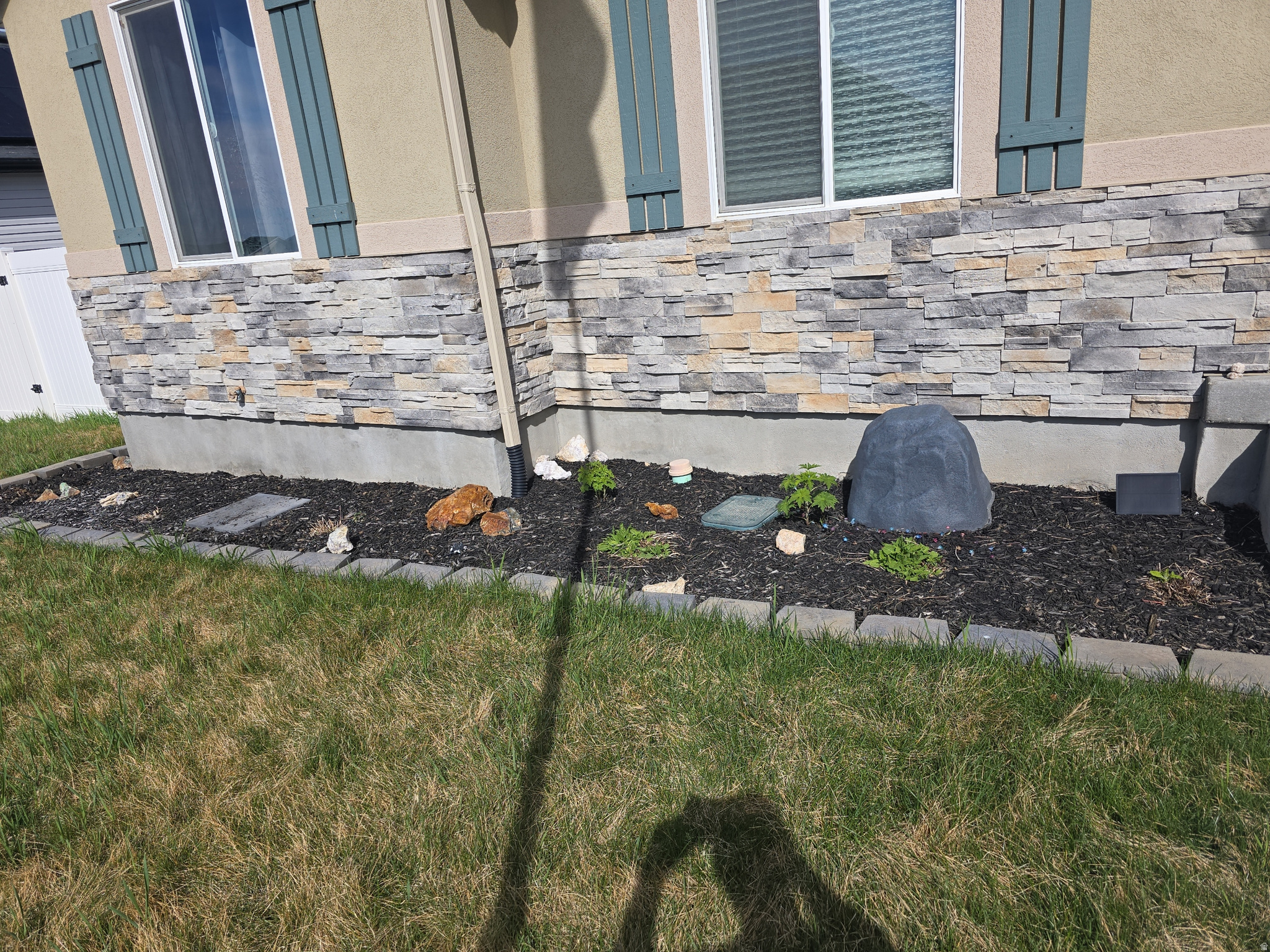 Exterior wall with stacked stone veneer, beige siding, decorative window shutters, and a landscaped bed featuring dark mulch and stone edging