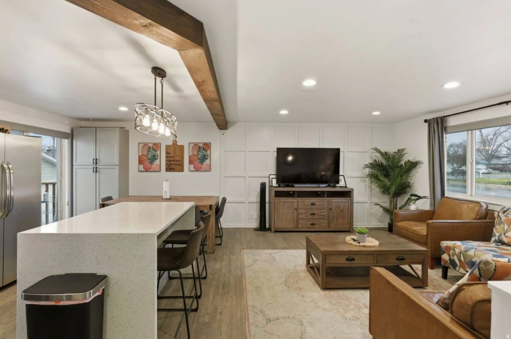 Open-concept living area featuring a kitchen island with seating, wood-finish flooring, a decorative ceiling beam, recessed lighting, and a window with natural light