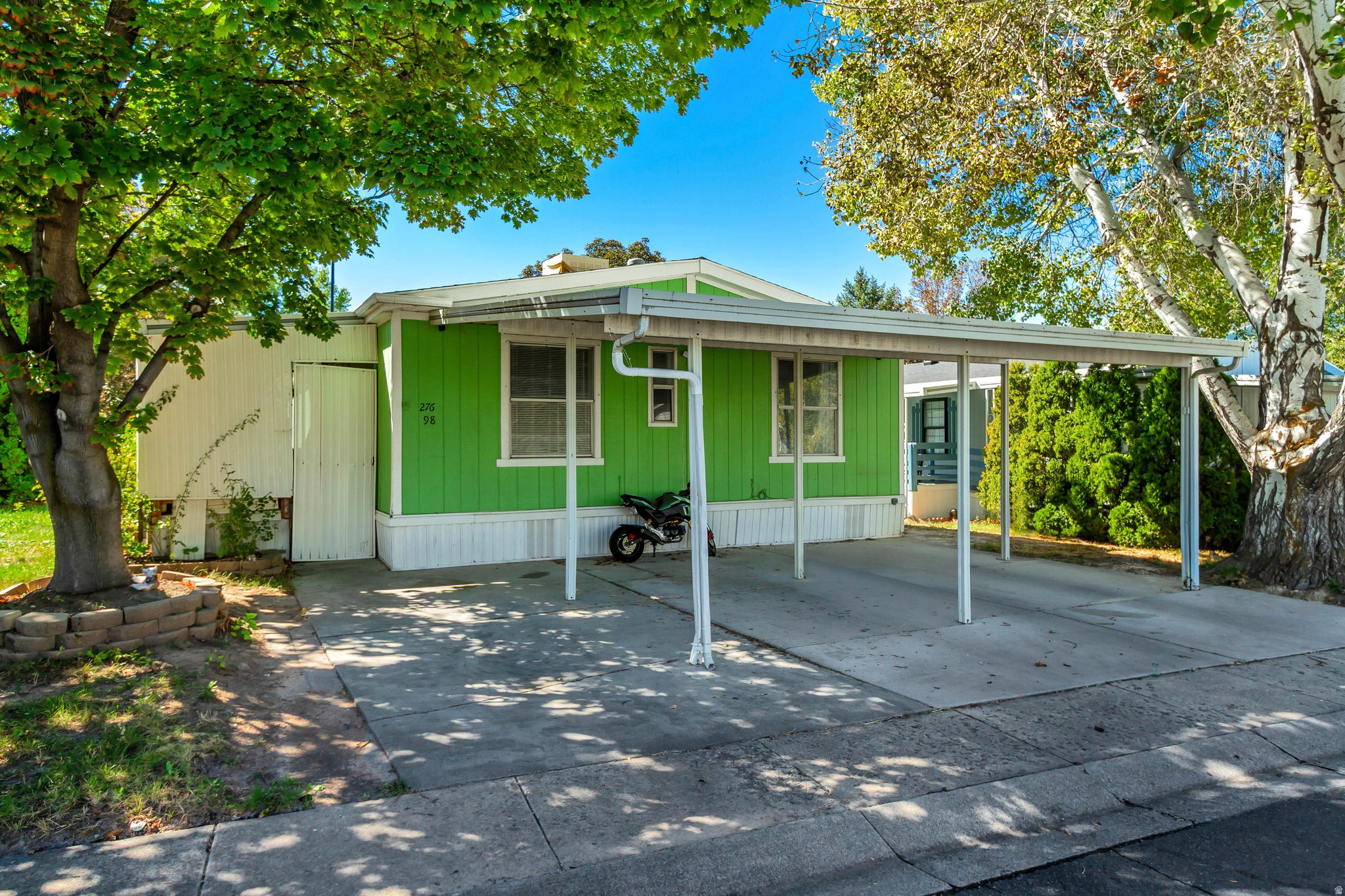 Green exterior siding with white trim, carport structure, paved driveway, mature trees, and an attached storage shed