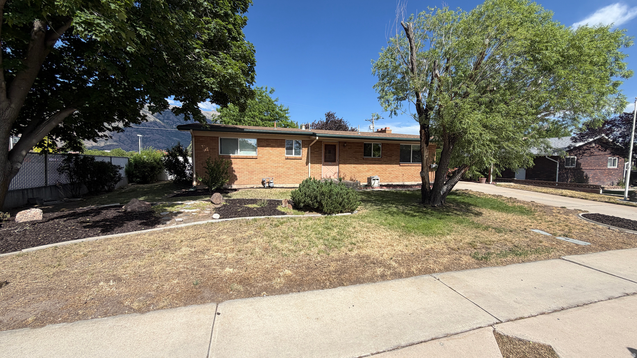 All Brick residence with a light gray roof, mature trees, a paved driveway, and a white vinyl fence