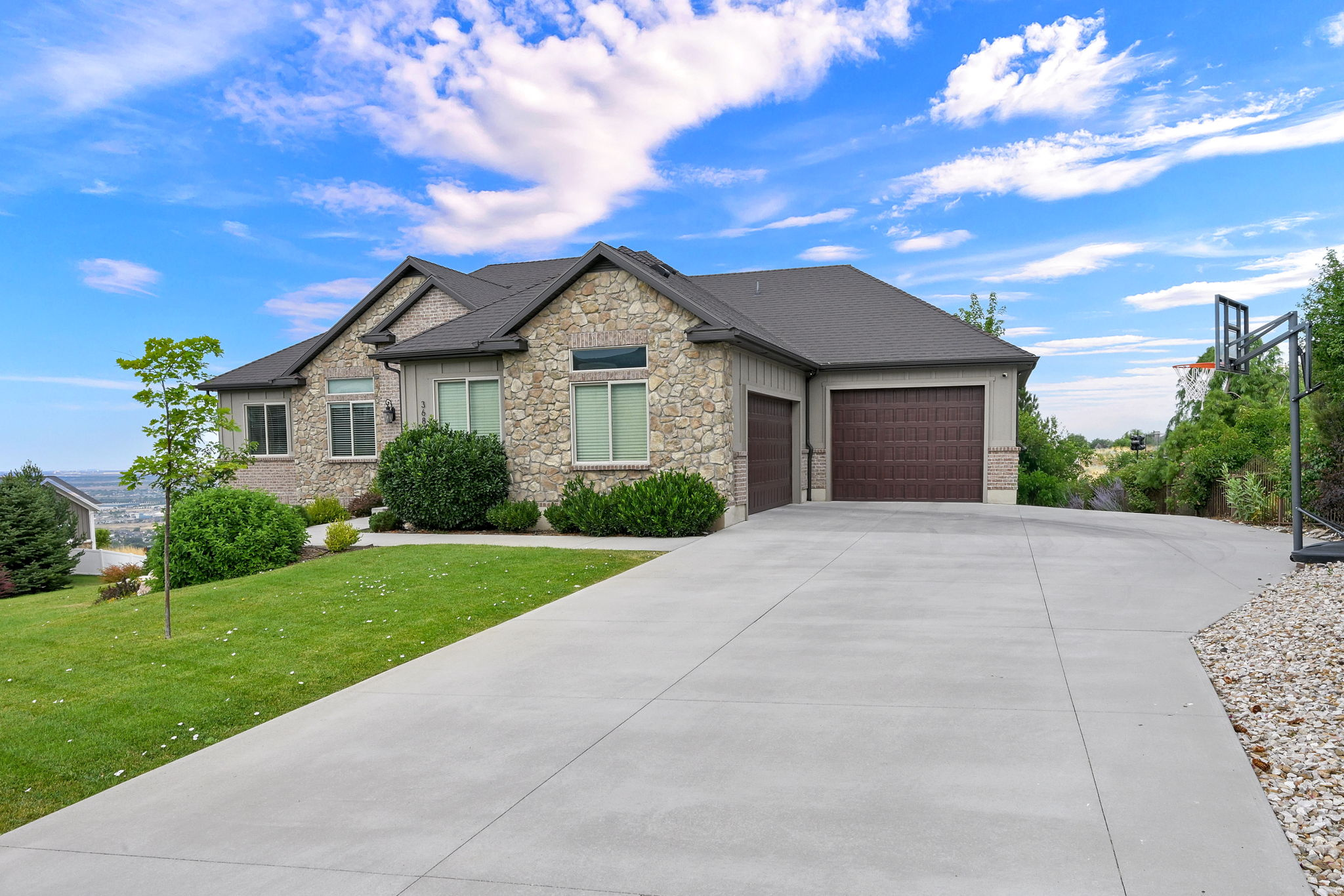 Stone and siding exterior featuring a multi-gable roofline, attached two-car garage with wood-finish doors, expansive concrete driveway, and established front landscaping