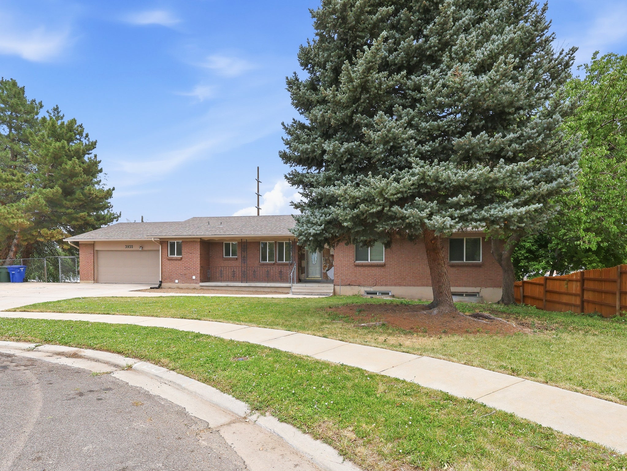 Brick exterior residence with a front-facing garage door, mature evergreen trees, a concrete driveway, a concrete sidewalk, and a fenced perimeter
