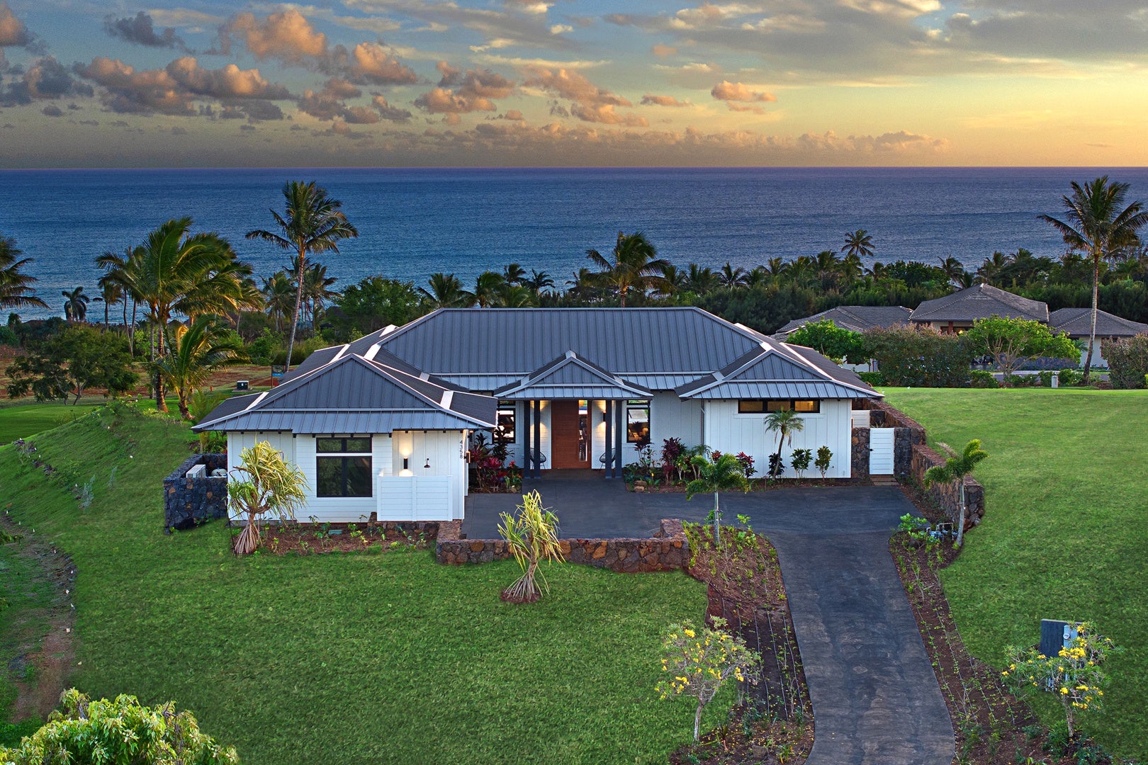 Modern white home with a gray metal roof overlooks the ocean from a landscaped lawn.