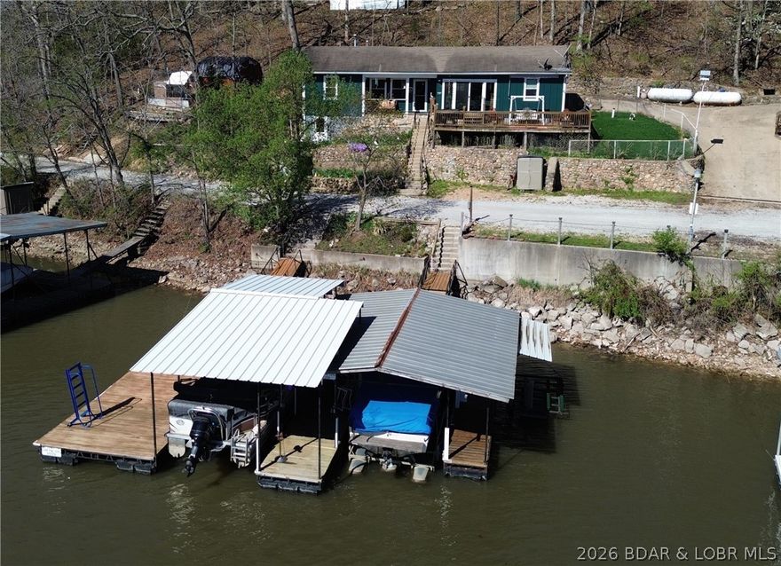 Lake of the Ozarks waterfront property featuring natural stone accents and a functional layout. The living room offers a full stone wall with fireplace and wood flooring. The primary bedroom is located off the living area with sliding barn door and includes a walk-in closet and attached bath with shower/tub combination. The kitchen and dining area open to the living space and provide access to a large deck overlooking the water. Kitchen features include stainless steel appliances, granite countertops, tile backsplash, and pantry. A laundry room with half bath is also on the main level. A second bedroom and partial unfinished basement offer additional space and storage.
This property includes a shared well, private septic system, shared driveway, storage building, and fenced kennel area. Waterfront features include a seawall and covered, two-well dock with boat and PWC lifts, and a swim platform, located in a cove setting at the 61MM with year-round deep water access. This property has a main channel view with fabulous sunrises! Come make this your lake home!