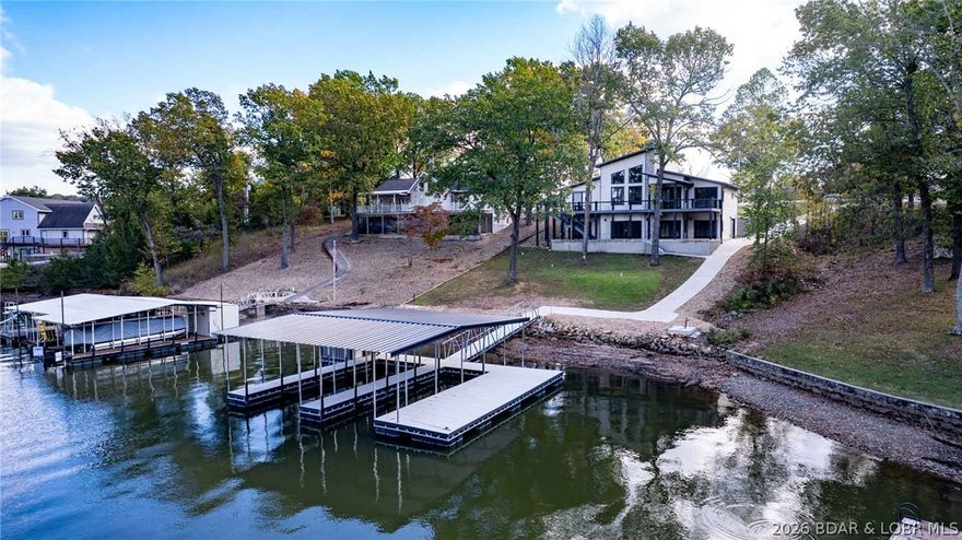 Discover lakefront luxury w/rare zero-step access to the dock—an increasingly hard-to-find feature at the LOZ. This architecturally designed residence blends modern sophistication w/effortless lakeside living. Inside, soaring vaulted ceilings, rich hickory flooring, & dramatic 8-foot sliding glass doors frame peaceful cove surroundings w/beautiful main-channel views. The chef’s kitchen anchors the home w/custom cabinetry, quartz surfaces, professional-grade appliances, & impressive 12-seat island built for entertaining. Two private en-suites offer spa-inspired baths & custom walk-in closets, creating true owner retreats. Premium features include heated lower-level floors, indoor/outdoor fireplaces, wine cellar, & spray foam insulation for exceptional comfort/efficiency. Over 2,000 sq ft of covered and open patio space extends the living experience outdoors. LP SmartSide siding, metal roof, and composite decking provide lasting beauty with minimal maintenance. Private dock with 12x34 & 10x34 slips, swim platform, storage shed included. New construction lakefront homes with this level of design and dock access are rarely available—an opportunity that won’t last long.