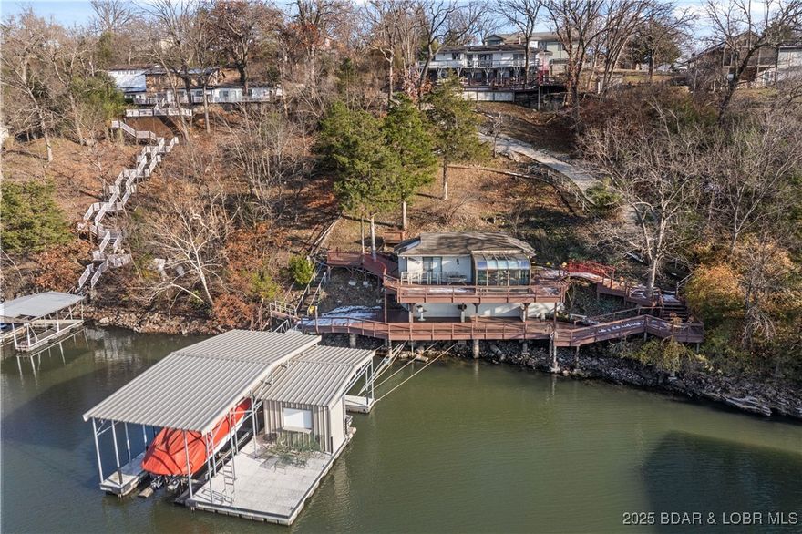 Don’t miss this incredible lakefront opportunity featuring a 5BD/4BA main home plus a 1BD/1BA guest house perched over the water on MM 17 in Lake Ozark! Offering big water views, a prime location by land and water, and nightly rental approval, this property is ideal for full-time living, multigenerational use, or investment. The one-of-a-kind guest house offers a large lakeside sunroom, full kitchen, living room, spacious bedroom, full bath, and extensive wraparound decking. Enjoy a newer 2-well concrete dock (12x36 + PWC slip) with a swim platform and storage/bar room with electric. The main home features two full kitchens (including a newly remodeled main kitchen), two living areas, two primary suites, two dining spaces, and a 2-car garage. Additional highlights include two wood-burning fireplaces, indoor/outdoor speakers, abundant decking (recently repaired and resealed), vaulted ceilings, newer roof and stucco on the guest house, flat driveway, main level living, and approx. 30’ of water depth. A new cart path to the dock adds easy access. Truly exceptional lakefront living—turn-key and ready to enjoy!
