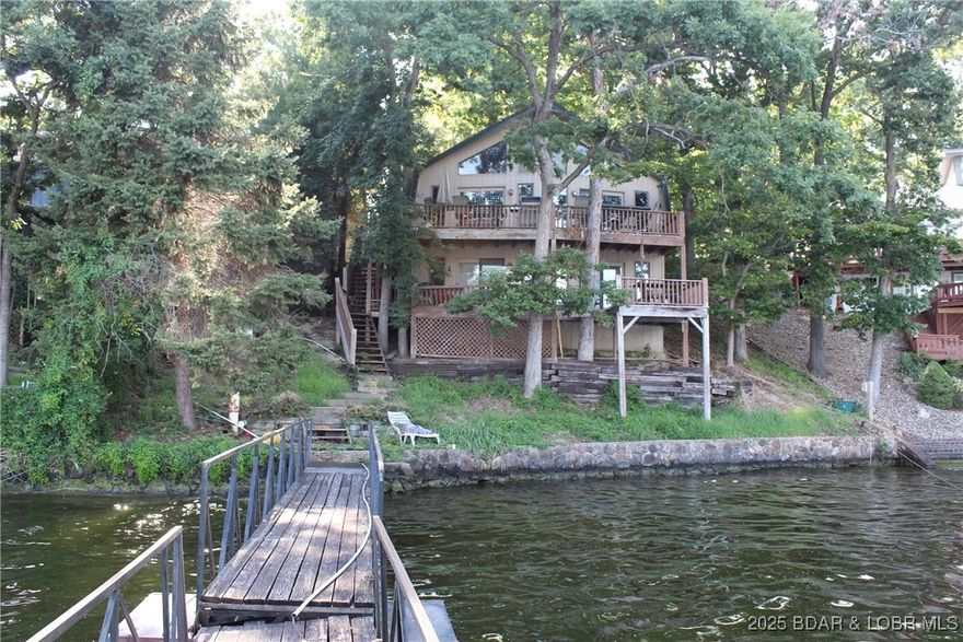 MOTIVATED SELLER! Lakefront Home with Stunning Views, Three-Well Dock & Endless Potential! Experience lake living with this 3-bedroom, 2-bathroom home featuring a bonus room and year-round views of the Lake of the Ozarks. Tucked away in a quiet inlet at the 7 MM of the Main Channel, it offers privacy while being just minutes from waterfront restaurants and attractions.  Inside, enjoy vaulted, hand-hewn wood ceilings, an open concept living area flowing into the kitchen, and two charming stained-glass windows. Two decks overlook the water, perfect for entertaining or relaxing. The lower level includes a second full kitchen, large living area with lake views, and fireplaces on both levels.  While updates are needed, the home provides a solid foundation to create your dream lake retreat, and renovation loan options may be available to help bring your vision to life. Outside, a three-well dock with swim platform and wet steps awaits, plus a one-car garage with extra storage underneath.  This property combines space, views, and location for the ultimate lake getaway.