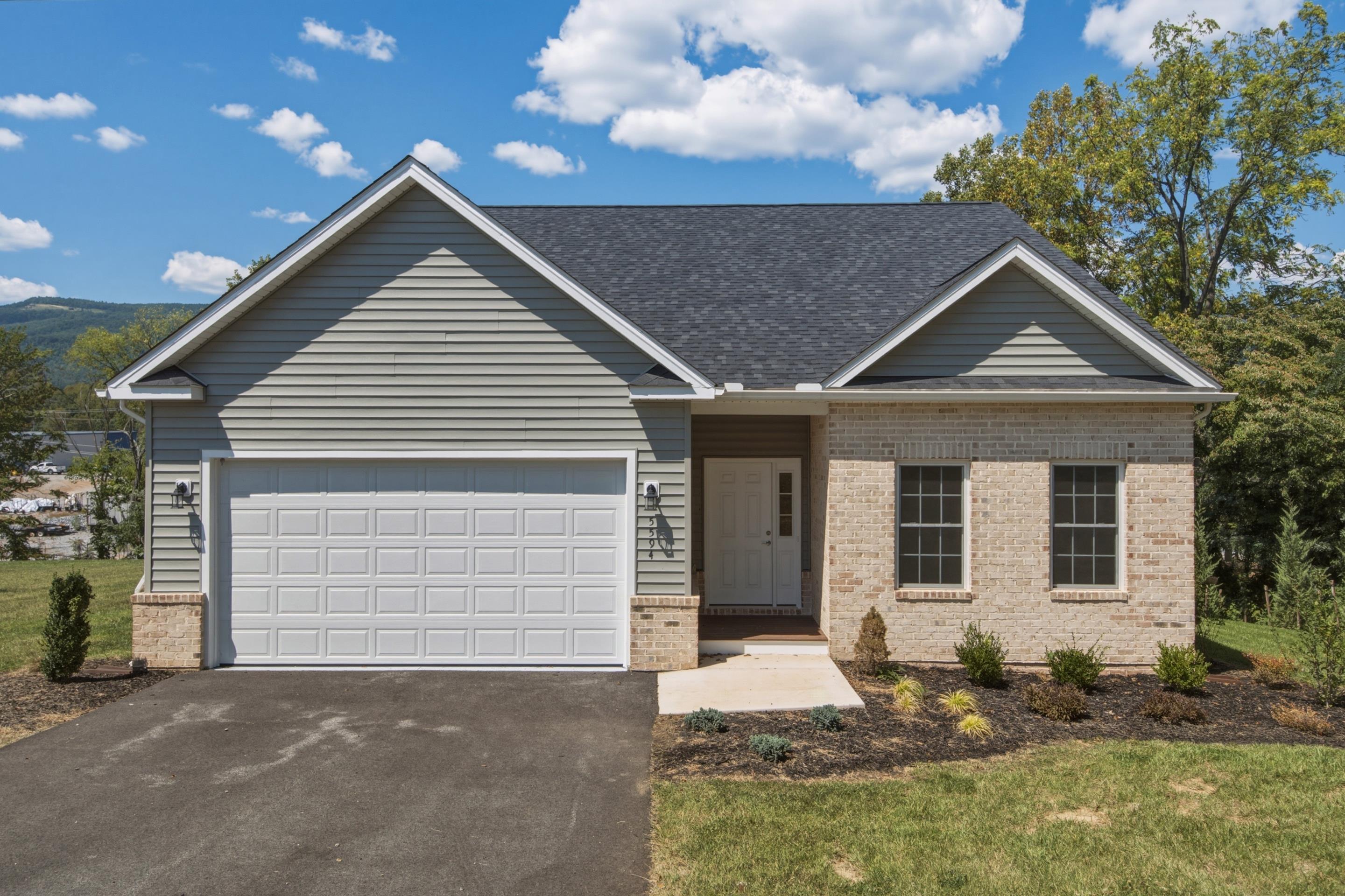 Single-story gray-and-brick house with a two-car garage, landscaped front beds, and wooded hills behind it.