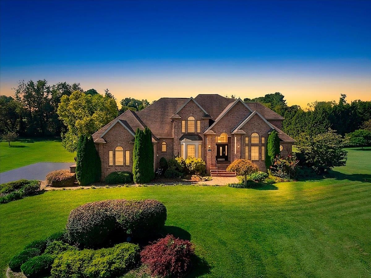 Brick house with multiple gables and illuminated windows, set behind a landscaped lawn at sunset.