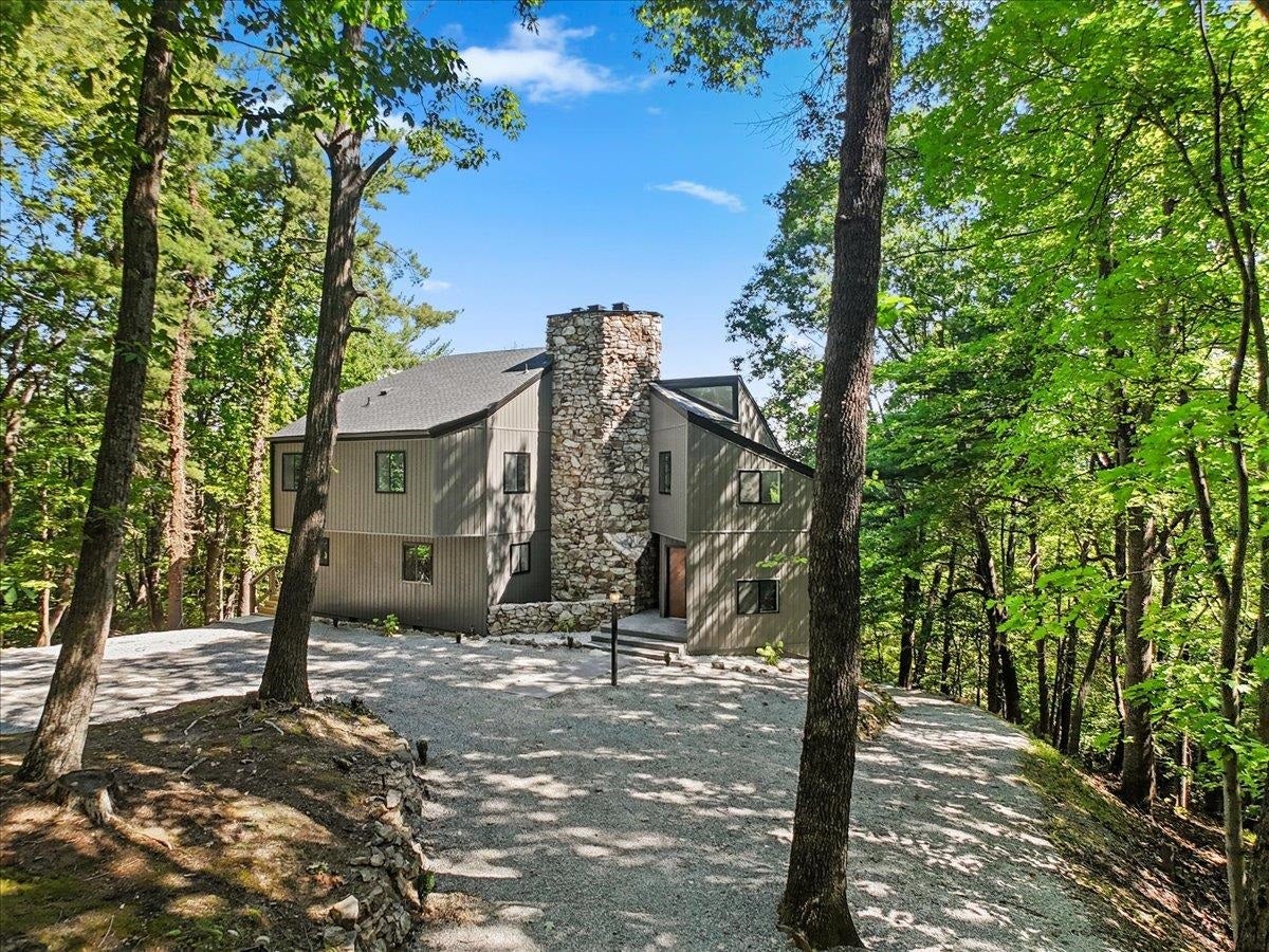Gray-sided home with a tall stone chimney surrounded by mature trees and a gravel driveway.