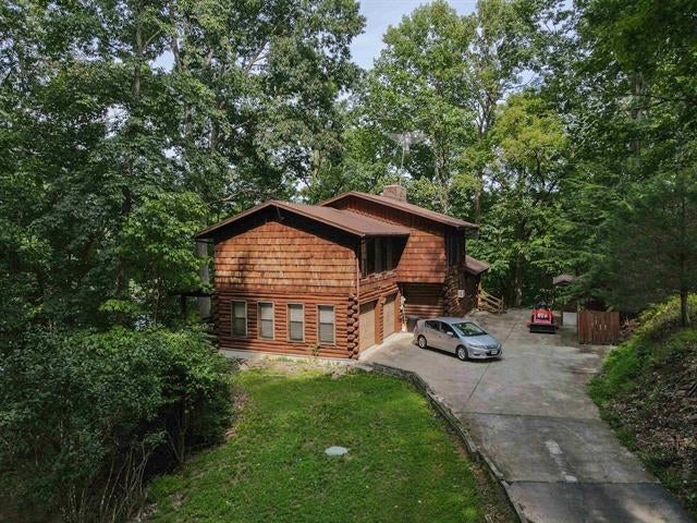 Wood-sided two-story home with a broad driveway, parked cars, and mature trees.