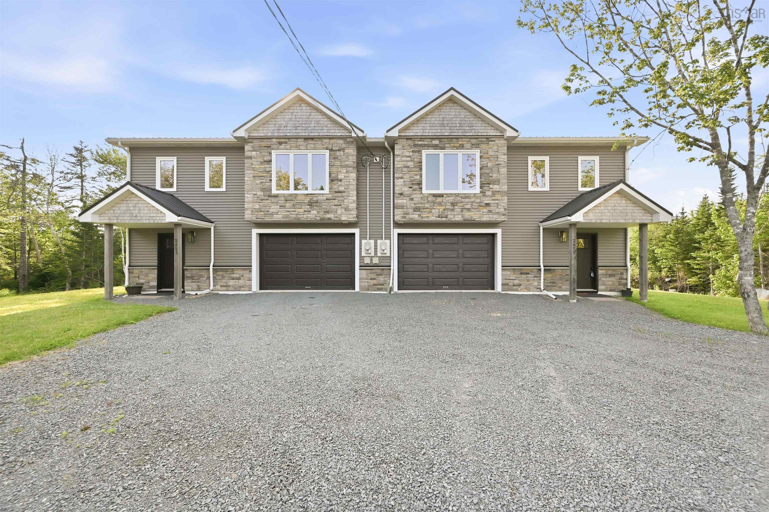 Duplex exterior featuring stone and siding finishes, individual gravel driveways, attached single-car garages, covered entryways, and a treed backdrop