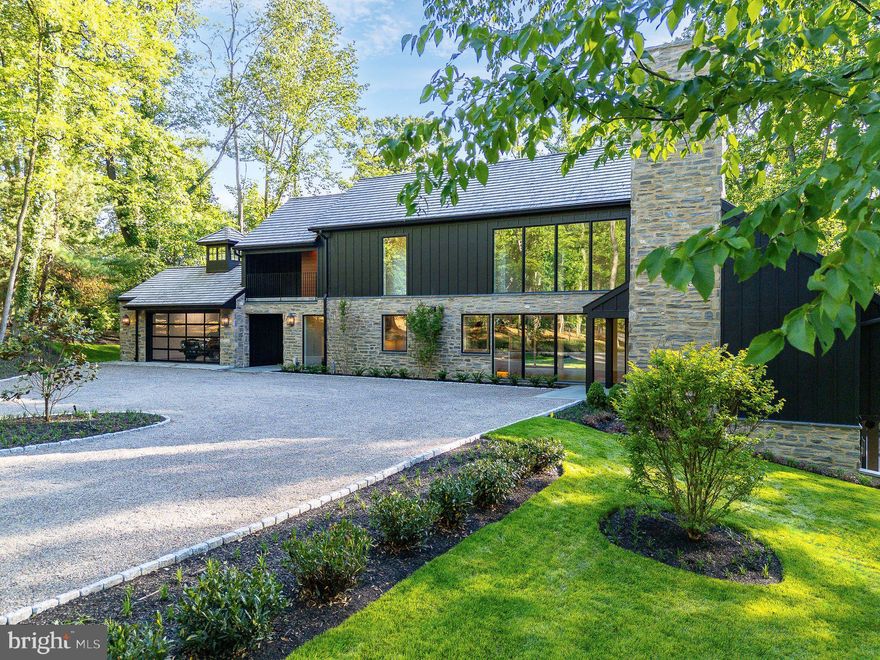 Welcome to 607 West Gravers Lane—a striking fusion of modern architecture and serene natural beauty. Designed by award-winning Blake Development Corporation, this newly built masterpiece is nestled on nearly two acres at the edge of the Wissahickon Valley in Fairmount Park, offering panoramic views that shift with the seasons—from lush greens in summer to a rich tapestry of fall foliage and winter textures.

A circular drive and landscaped island provide a gracious welcome. The home’s exterior artfully blends natural stone, board-and-batten siding, and varied rooflines for a look that’s both timeless and contemporary. Bluestone entryways hint at the refined design within, where modern elegance meets warm, livable luxury. Spanning 6,100+ square feet across three levels, the home features 5 bedrooms, 4 full and 2 half baths.

The dramatic entry foyer, with soaring ceilings, a statement pendant, and a picture window, sets the tone. Wide white oak floors and coffered ceilings create a clean-lined, airy sophistication throughout. In the main living room, exposed beams and a two-story natural stone gas fireplace anchor the open-concept space, which includes a stylish wet bar and adjacent lounge with herringbone-patterned floors—perfect for entertaining.

The chef’s kitchen is a true showstopper with Sub-Zero, Wolf, and Bosch appliances, waterfall countertops, and custom cabinetry. A flagstone terrace off the kitchen is ideal for coffee or cocktails amidst the trees. The main level also includes a private ensuite bedroom, office or optional sixth bedroom, powder room, and laundry with built-ins.

Upstairs, an open-tread staircase leads to a dramatic overlook and two bedrooms, one with a charming loft, plus a hall bath. The crown jewel is the primary suite: gas fireplace, boutique-style dressing room, and a spa-like bath with a sculptural soaking tub, dual-entry doorless shower, and heated marble floors for ultimate comfort.

The lower level continues the experience with a glass-enclosed wine room featuring stone walls and flagstone floors, a game/media room, gym, ensuite bedroom, laundry, and ample storage.
Multiple outdoor terraces invite al fresco dining and tranquil moments, surrounded by a professionally designed, mature landscape that enhances privacy and complements the Wissahickon setting. Select customizations are still available for the discerning buyer.

Located in one of Chestnut Hill’s most coveted enclaves, 607 West Gravers Lane offers an extraordinary opportunity to enjoy modern luxury harmonized with nature.