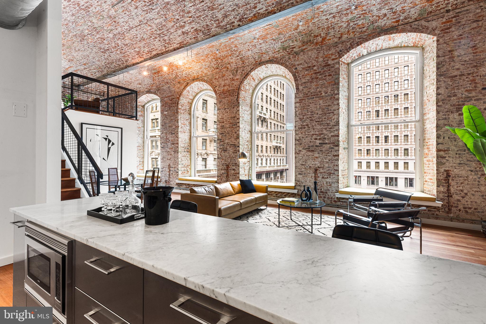 Residence No. 300 at The White Building is a quintessential corner loft situated in one of Philadelphia’s favorite neighborhoods and within the McCall School Catchment. Built in the late 1800s, this former factory building has since been converted to luxury lofts that thoughtfully blend pre-war architecture with refined living space; No. 300 may be the best expression of this. With details like preserved barrel-rolled ceilings, soaring brick walls, arched windows, and steel factory doors, original character is found at every turn. A long hallway greets at the entryway of the property. Here, overhead gallery lighting illuminates the first of many exposed brick walls, which can be left barren or serve as a gallery wall lining the walkway to the living room. Opposite the wall leading in are two coat closets and a cutout for an entryway table. As you advance to the open living area, relics from another age are on full display. 20-foot barrel-vaulted ceilings and oversized arched windows with sill benches frame the northern wall of the grand room that looks onto the recently completed Jefferson Plaza. The high-end kitchen is steps away and features striking Pedini cabinets and drawers, a Carrera marble countertop with a drop-down breakfast bar, and Bosch and SubZero appliances. The continuity between the living room, kitchen, and dining area makes hosting a breeze. A lofted den sits atop the open front room; this is the perfect lounge, record, or media room to retreat to in the evening. There is additional space for an at-home office, as well a storage nook under the stairwell that could be converted to pantry shelving. Continuing on, a hallway between the living room and primary bedroom contains a laundry closet with Bosch stackable appliances as well as a tiled full bathroom complete with a soaking tub and overhead shower, deep-sink vanity, frosted cabinet, and chrome Brizo fixtures. The primary suite impresses with its glass and steel factory doors that line the entryway closets leading into the bedroom. King-sized furnishings fit this room well, and there is additional space for a bedroom lounge or a large chest of drawers near the west-facing window. The en-suite bathroom offers a glass-framed shower with chrome hardware, a soaking tub with dual faucets, a large vanity, and tiled flooring and walls. A second bedroom is accessed via the living room. With two west-facing windows and a shelved closet, this room enjoys a generous amount of daylight and is steps away from the full hallway bathroom. No. 300 additionally offers 200 square feet of in-unit attic storage with pull-down stairs. The White Building features a sleek lobby with secure doors and elevators, a mail room, and a mezzanine-level gym. ** A credit towards monthly parking at a nearby garage is offered. ** In addition to living in one of Philadelphia’s favorite condominiums, building residents are steps from some of the city’s best restaurants, shops, gyms, and more. We look forward to showing you this inspiring space and the lifestyle that comes with living in a boutique community in the center of it all! Disclaimer: Listing broker and salespersons do not guarantee the estimated square footage or tax data, which should be independently verified by the buyer and buyer’s agent/s. The 1657 square feet estimate is derived from floorplan measurements, including the lofted den in the calculation, but not the attic space.