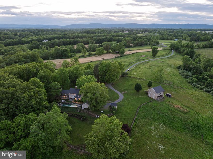 Mayapple Farm | This is an incredible opportunity to live minutes from Middleburg on an extraordinarily unique property.   The house is rich in character, known as the Nathan Ayer House, and was built in 1790 in Preston City, Connecticut, dismantled, and moved to Virginia hunt country.   The quality and authenticity throughout the home is remarkable – the charming floorboards, 9 stone fireplaces, custom woodworking, original hardware, and exposed beams only begin to describe the features of this home.  The kitchen, primary bathroom, and guest bathroom have been tastefully renovated by the current owner with quartzite counters, custom cabinetry, and high-end finishes.  There is an inviting pool, terraces, and lush perennial gardens off the rear of the house, all surrounded by stately dry stacked stone walls.  The bank barn dates back to the late 19th century and is constructed of hand-hewn oak and poplar beams and fieldstone, originally built in Leesburg, Virginia.  The stone barn offers endless possibilities for entertaining, creative workspace, and, of course, horses.  The home is heated and cooled by a geothermal system with seven separate zones.  The property has frontage and access to Goose Creek and provides ultimate privacy.