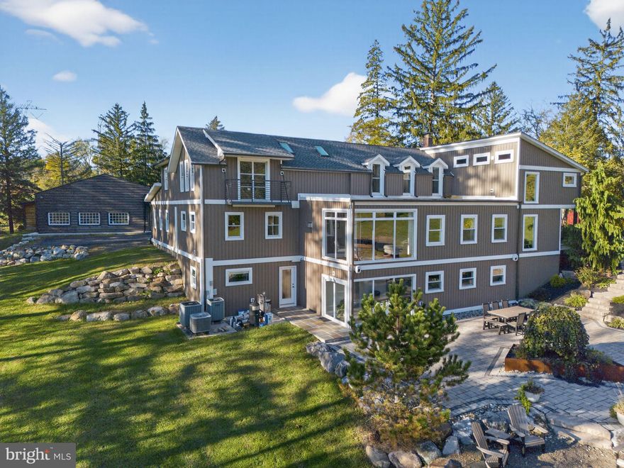 Set on over 9 private acres with sweeping countryside views in Lower Milford Township, this exceptional property blends the historic character of an 1880s stone farmhouse with modern design and thoughtfully curated spaces throughout. Portions of the home’s original fieldstone have been carefully exposed and highlighted, creating a striking mix of historic charm, modern farmhouse style, and industrial inspired accents. A private driveway leads to the home, where the main living spaces are designed to showcase the landscape beyond. The living room is truly remarkable, featuring floor to ceiling windows that frame panoramic views of the property and fill the space with natural light. A gas fireplace anchors the room, while the scale of the space creates an impressive setting for both everyday living and entertaining. The spacious chef’s kitchen has been thoughtfully updated and serves as the heart of the home. Designed for both everyday living and gathering, it features stainless steel built in appliances, a Wolf six burner gas range, double ovens, and an oversized center island topped with a striking black walnut countertop, complemented by custom metal detailing and statement lighting above. Natural stone accent walls, a built in bench, and a spacious pantry with sliding barn doors add both warmth and character to the space. The dining room continues the home’s distinctive design with exposed wood ceiling beams and natural stone walls, creating a space that feels both timeless and inviting. A separate den or study offers a cozy secondary living space with a wood burning fireplace, wainscoting, and additional custom metal accents. The main level also features an exceptional mudroom and laundry area located just off the garage, offering abundant space, built in bench seating, and a convenient dog wash station, making it as functional as it is impressive. A half bath completes this level. Upstairs, the primary suite captures peaceful views of the property’s pond and creek. The spacious en suite bath features a soaking tub, double vanity, skylights, and a walk in closet. Four additional bedrooms are located along a bright hallway and each features cathedral ceilings, creating airy, light filled spaces. A full bathroom with a walk in shower and skylight serves the additional bedrooms on this level. The finished walk out lower level adds even more flexibility with a large open recreation area that leads directly to the backyard patio, creating an ideal space for a game room, media room, or additional entertaining area. This level also includes a full bath and a private office. Outdoor living is just as impressive as the interior. The backyard has been designed for gathering and relaxation with a salt water in ground pool, built in stainless steel outdoor kitchen, firepit area, and stone patio overlooking the property’s open landscape. A treehouse and chicken coop add to the charm of the grounds while maintaining the privacy and space that nine acres provide. In addition to the oversized attached two car garage with nine foot doors, epoxy flooring, and direct access into the kitchen, the property also includes a large detached outbuilding featuring a finished 33 x 20 gym area, an attached 38 x 50 garage with two 11 foot doors and built in workbench, and an additional14×36 area off the garage with a separate entrance—perfect for storing your farm and lawn equipment. A rare opportunity to own a property that combines acreage, privacy, architectural character, and thoughtfully designed living spaces both inside and out.