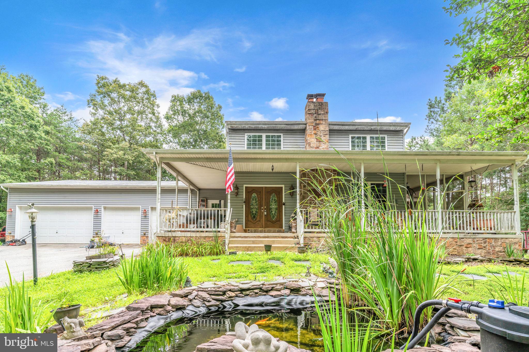 This spacious Colonial-style home sits on 4 wooded and private acres and offers over 4,000 square feet of living space. The oversized attached 4-car garage—complete with heavy-up electric, workbench, and multiple bay doors—makes this property a dream come true for hobbyists, car enthusiasts, or anyone needing serious workshop space. While the home could benefit from some updating, it is priced to reflect the condition and offers incredible potential. As you approach, you’ll be greeted by a wide covered front porch overlooking a peaceful karaoke pond—ideal for morning coffee or unwinding in the evening. Step through the double front doors into an expansive great room featuring vaulted ceilings, a floor-to-ceiling stone fireplace with pellet stove insert, recessed lighting, dual ceiling fans, custom log mantle, and abundant windows that fill the space with natural light. Hardwood floors flow throughout the main level.
The main level also offers a private office with LPV flooring, recessed lights, and ceiling fan, a main-level bedroom with double closet, and a full bathroom with marble vanity and tub/shower combo. A large screened-in porch—connected to the front wraparound porch—sits off the great room and is wired for a hot tub. It’s ready for your finishing touches with new ceiling fans and screens. The kitchen area leads to a mudroom-style laundry room with extra cabinetry, laundry sink, fluorescent lighting, and newer front-load washer and dryer. The upper-level owner’s suite is generously sized and features a sitting area, massive Texas-sized walk-in closet, and a full ensuite bath. New sliding doors off the suite provide access to private balcony. Downstairs, the fully finished basement includes a spacious recreation room, additional bedroom, and full bath. The basement also offers a separate entrance via new doors and has a sump pump, a second pellet stove (not yet installed, tile and ductwork convey), and extra materials including drywall and brick pavers that are included in the sale.
The oversized four-car garage includes a large two-car bay door, two single doors—one with extra clearance for trucks—and is fully equipped with shelving, a workbench, and two brand-new garage door openers ready for installation. Additional highlights include dual-zone heat pump systems, a water conditioning system, and a new well pump installed in 2024. There is also a designated forest conservation area within the 4-acre lot. The 30x18 wooden deck with pergola offers the perfect space for outdoor entertaining, with steps leading to a brick patio and former pool area—now ready for repurposing. This is a rare opportunity to own a private, spacious Colonial with serious garage space and endless potential—sold as-is and ready for your custom touches.