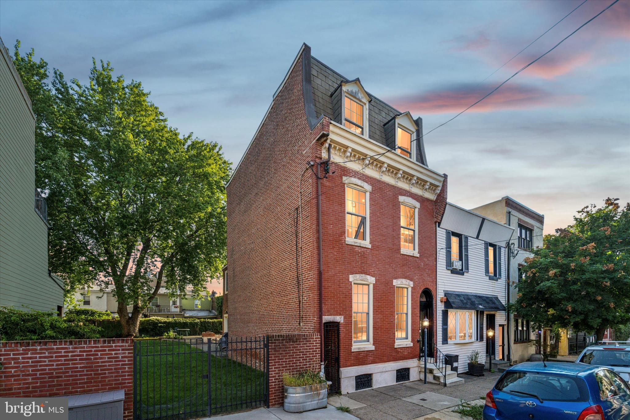 Step into a rare Philadelphia treasure—an elegant 1875 Second Empire rowhouse with Italianate detailing, set on an expansive lot in East Kensington that only 1% of single homes in the area can boast. The mansard roof, decorative cornice, arched windows, and preserved Victorian touches like stained glass, antique lighting, marble fireplace, wood trim, and banisters reflect a timeless grandeur, while modern updates make it ready for today. Inside, original wood floors lead you through a spacious layout. The double living room gives you flexibility—create a formal dining space, a quiet reading nook, or a home office in one section, while the larger area easily accommodates a wrap-around sofa for cozy movie nights or lively conversations. The open kitchen and dining area are bathed in light, with 2 sets of double doors opening to the yard, creating a seamless indoor-outdoor flow. Step outside to a backyard oasis unlike anything in the city—decking with built-in fire pit and planters, stone patio, and a yard large enough to host a wedding, family reunion, or your personal pool. Beautiful brick walls and lush vines enclose the space, offering both privacy and charm. A chef’s dream kitchen features granite countertops, stainless steel appliances, a professional 6-burner Premier Pro Series gas range, workstation, recessed lighting, water filtration, and exposed brick that rises up the entire right side of the house. The second floor offers three spacious bedrooms plus a den, all with endless layout options—bedrooms, playroom, guest rooms, dressing room, or home offices. The massive front bedroom is especially noteworthy. Upstairs, the third-floor primary suite feels like a retreat, complete with two double closets, skylit spa bath with Jacuzzi tub, and the potential for a roof deck with skyline views. The full-sized basement features a finished ceiling and recessed lights, making it perfect for a playroom, gym, or media room, as well as laundry, storage, and a utility tub. 8-minute walk to the Berks El Station and close to all the neighborhood favorites on Front and Frankford Streets, plus Riverwards Produce, this home combines historic character, modern comfort, and unmatched outdoor space. A true one-of-a-kind opportunity in East Kensington.