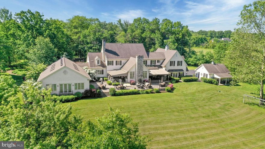 Set on an enchanting 30 acres, down a winding driveway bordered by tall trees on a quiet country road in Upper Bucks County, sits Four Seasons Manor. Surrounded by wide lawns and lush landscaping, this stately custom home is designed to take full advantage of its surroundings, with almost 10,000 finished square feet inside, and more than 3,000 square feet of patios, porches and terraces. The circular driveway leads to the handsome formal entry, with a deep covered porch that features a barrel ceiling over the front door with its beveled glass window and sidelites. Inside, the home’s open floor plan is a counterpoint between romantic design themes and functional practicality. Warm hardwood floors, high ceilings, detailed moldings, and abundant windows make a dramatic first impression. While the exterior suggests a mood of formality, the openness of the interior, with spaces that flow gracefully from one area to the next, add a perfect balance of casual informality. Located at the front of the home, the office takes full advantage of its space with multiple desks, built-in storage, and even a moveable worktable. The formal dining room is enhanced by bringing the outdoors in with French doors to the front porch, an architectural theme that also encourages graceful entertaining. In the kitchen, cherry cabinets, granite countertops and Travertine backsplashes create a harmonious contrast to the ever-changing beauty of the natural surroundings. Paneled appliances, gas cooking, and abundant prep and storage space will please even the most discerning of home cooks. From here, the floor plans flows into a large breakfast area, and then to the living room with its coffered ceiling, gas fireplace with marble surround, and French doors to the rear patio. The family room is a magnificent, beautifully proportioned “great room” with a two-story vaulted ceiling, complemented by an exposed stone wall with gas fireplace, and romantic interior Juliet balcony on the upper level that looks down from the primary suite. A bedroom and full bath on this level add flexible space for guests or extended family. Two sets of stairs lead to the second floor, where the expansive primary suite is a welcome respite from the outside world. With a tray ceiling and large walk-in closet, the bedroom opens to a sitting room and en suite bath with dual sinks, air-jet tub, and multiple closets. Two large bedrooms share a hall bath, while a jack-and-jill bath separates two additional bedrooms that each have their own loft space accessible by ladder. Another suite on the third floor has its own walk-in closet as well as storage space. The finished lower level includes a recreation room, kitchenette, bar area, full bath and sauna. The piece de resistance is the massive indoor pool, designed to integrate with the main house for year-round enjoyment. Situated in its own wing, the room is an architectural marvel featuring a vaulted ceiling with skylights, walls of windows, and French doors to the yard. The saltwater pool includes three underwater corner seats, a retractable cover, and a diving board. The nearby hot tub seats 10, while a fully equipped wet bar makes it easy to enjoy the space with friends. By day, the home exudes formal elegance. By night it’s a theater for bold drama around the terraces. Entertaining is a joy when guests can savor the home’s exterior beauty, while enjoying glimpses of the interior. Flagstone and paver patios are accessible from nearly every room of the house, with awnings for shade and a wood-burning fireplace for chilly evenings. Nearby, the detached garage provides covered parking as well as storage, along with garden and equipment sheds. A nearby pond with charming curved footbridge features a waterfall that runs off its own filtration system. Solar panels and an automatic generator ensure the property runs efficiently and without interruption year round.