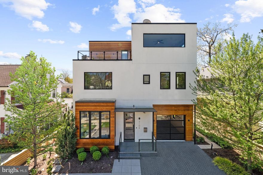 Steps away from urban Downtown Bethesda is 5106 Wilson Lane, set back on a quiet tree-lined, slow-moving portion of the famed street. With its right angles and mix of wood siding, stucco, and glass, the home’s contemporary design is a stark departure from more traditional styles, symbolizing the Bethesda of the future. The floor plan comprises nearly 6,000 finished square feet of premium living space spread across four open-concept levels.

The entire main level nearly consists of a single unbroken space encompassing living, dining, and kitchen areas. The former boasts a modern linear fireplace with accordion-style doors, fully opening up the space to indoor/outdoor living, allowing the rear patio to seamlessly extend the living space. The latter is stunning, with its oversized natural stone waterfall center island, minimalist European cabinetry, and a full suite of SubZero-Wolf appliances, including a column wine chiller. It opens up to an inviting screened-in porch ideal for dining al fresco.

Ascending the light-filled wood and glass staircase reveals the first upper level, home to a spectacular primary suite. Its amenities are boundless — a separate sitting room with fireplace, custom walk-in closet with extensive built-ins, and spa-inspired bath with twin vanities, freestanding soaking tub, and jetted shower combine to make the space a total escape. The two secondary bedrooms on this level are both ensuites.
On the top level are three additional bedrooms, two of which share a large buddy bath with separate vanities. This level’s party piece is the rooftop deck which offers stunning views of the downtown skyline, a truly unique feature.

For the entertainer, the lower level boasts every attraction—from a recreation room to a wet bar, fitness studio, temperature-controlled wine grotto, professional home cinema, and additional full bath—with each element executed to perfection. The entertainment does not stop once out back, as the rear patio steps down to an oversized hot tub nestled in a private, cozy corner. On the opposite side of the rear lawn is the fully-equipped outdoor kitchen, whose adjacent roaring fireplace is the ideal gathering spot for s’mores or stargazing on a brisk night.

With its walkable location, the home belongs to one of the nation’s finest school clusters and is easily accessible to Bethesda, Chevy Chase, DC, and major commuter routes. Combining this unbeatable location with all of the square footage, amenities, and designer touches found within the home, 5106 Wilson Lane is for the most discerning buyer.