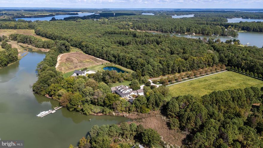 Set along the peaceful waters of Caulk Cove, this extraordinary waterfront estate offers luxury, privacy and natural beauty across more than 10 acres. The property features open farm fields and professionally landscaped, park-like grounds, with a tree-lined driveway creating a breathtaking approach to the residence. Built in 2013, the custom-designed home encompasses nearly 8,500 square feet of thoughtfully curated living space, positioned to capture sweeping waterfront views. A gracious front porch welcomes you into a grand foyer, setting the tone for the exceptional craftsmanship found throughout. The main level offers soaring 12-foot ceilings, rich hardwood floors, expansive windows and doors and custom millwork including crown molding, coffered and tray ceilings and elegant board-and-batten detailing. At the heart of the home, the living room serves as a remarkable gathering space with a dramatic floor-to-ceiling stone fireplace, custom built-ins, coffered ceilings, a wet bar and access to the rear porch. The kitchen is a stunning focal point, featuring a diamond coffered ceiling, custom handmade walnut cabinetry, two oversized islands, white and leathered granite countertops, and a Wolf six-burner, dual-oven range. A fully equipped butler’s pantry offers extensive cabinetry, generous counter space, a secondary refrigerator, dual dishwashers, a wine refrigerator and an electric oven. The open dining area flows seamlessly between the kitchen and living room. The primary suite is a private retreat with a tray ceiling, wood-burning fireplace, expansive walk-in closet with custom built-ins and vanity and a luxurious bath featuring heated floors, a jetted soaking tub, separate vanities, an open tile-and-glass shower and a private water closet. The suite is completed by a private sitting room with fireplace and wet bar, along with a private screened porch overlooking the gardens and serene waterfront. Four additional ensuite bedrooms each offer unique and luxurious bath designs. A well-appointed laundry room with ample cabinetry and generous counter space enhances everyday convenience and leads directly to a functional mudroom with access to the attached two-car garage. The second level features 9-foot ceilings and is anchored by a spacious family room with a stone hearth wood-burning fireplace and built-in window seating, flowing into a billiards area with wet bar, creating an ideal space for entertaining. The sixth bedroom currently includes custom built-in bunk beds but was originally designed as a home office and offers flexibility to be restored to its original use. The home’s exceptional design extends seamlessly outdoors, with a spacious screened porch leading to an open-air porch with a wood-burning fireplace and outdoor entertainment setup. Bluestone walkways and patios lead to a 20’ x 40’ lagoon-style Gunite pool with a waterfall feature, an outdoor kitchen and a waterside fire pit offering stunning sunset views over the cove. The property also includes a charming one-bedroom guest house with a luxury kitchen, open living area with fireplace and screened porch. A private dock with approximately 6’ MLW offers two boat slips, a floating dock and access to waterfront dining and premier fishing throughout the Chesapeake Bay and surrounding tributaries. Additional highlights to the home include a greenhouse with raised garden beds, a fenced garden area, professional landscaping with accent lighting, and a full irrigation system.  This exceptional estate is truly one-of-a-kind!