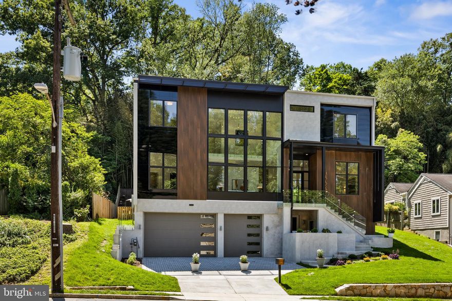 Set along Military Road and surrounded by North Arlington’s most established and prestigious neighborhoods, this modern estate offers a rare balance of architectural distinction and effortless access. Minutes to downtown Washington, Georgetown, Tysons, Clarendon, Amazon HQ2, and with convenient routes to Reagan National and Dulles International airports, the location supports an executive lifestyle that values both global reach and quiet residential living. Nearby trails and parkland add daily access to nature, reinforcing balance, wellness, and ease. Arrival begins on the main level through an 11-foot glass entry, opening to a composed sequence of formal spaces designed for presence and scale. At the front of the home, a grand living room rises to dramatic 25-foot ceilings—an exceptionally rare volume in Arlington—framed by glass railings above, modern lighting, and a full-height wall of glass. The formal dining room, also positioned at the front, stretches nearly 20 feet in length and comfortably hosts gatherings of fourteen or more, offering entertaining capacity seldom found at this level. Completing the formal front is an executive study of uncommon size, suitable for conference seating or dual workstations, with refined finishes and visual connection through the home to the pool beyond. As the home unfolds toward the rear, formal spaces transition seamlessly into relaxed yet equally impressive living. The great room, kitchen, and breakfast area open fully to the outdoors, where the saltwater pool becomes a central visual element—visible from front to back—linking the home’s formal and informal areas while filling the interior with natural light. European Porcelanosa cabinetry, Miele appliances, a waterfall quartz island, and a walk-in pantry with over 120 feet of storage anchor the kitchen with clarity and precision, while modern beams and a statement fireplace define the great room. Upstairs, glass railings overlook the living room below, reinforcing the home’s vertical openness. The owner’s suite is a private retreat with vaulted ceilings, skylights, uninterrupted pool views, and a discreet glass-wrapped balcony. Dual walk-in closets—together nearing 400 square feet—are thoughtfully separated and fully built out with illuminated cabinetry and custom storage. The spa-like bath features large-format stone, a glass-enclosed wet room, and a soaking tub designed for daily restoration. The lower level is unexpectedly light-filled and serves as the primary entry. Direct access from the oversized three-car garage—with tall doors suited for large SUVs—leads to a refined mudroom and storage spaces of exceptional scale. Additional driveway parking for three vehicles and ample street parking enhance everyday convenience. This level also includes a private bedroom suite, fitness room with glass entry, and direct walk-up access to the pool. Throughout, sanded-in-place white oak floors, wide staircases, and modern glass details create continuity and calm. Beneath the surface, construction quality quietly distinguishes the home: thicker engineered floor systems reduce vibration, 2x6 load-bearing walls enhance insulation and acoustic comfort, and thoughtfully engineered assemblies support durability and wellness. This is a residence for buyers who value discretion, access, and enduring craftsmanship.