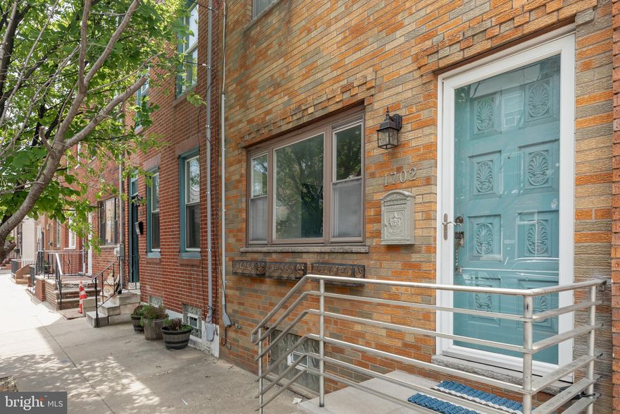 Finally, the home you have been waiting for is here. Come see this stylish and highly functional home just a block from East Passyunk Avenue, one block south of The Fountain, in the heart of the neighborhood.

Step up to this historic row home, refaced in mid-century brick and augmented by a 1950s front door and aluminum porch railing. Entering through the front door lands you in a tastefully tiled foyer offering privacy and separation from the spacious living area. The foyer wall features a beautiful transom window, while the reverse side provides the perfect place for your flat screen, art, or projections.

Beyond the foyer, the space opens into the uninterrupted flow of the living, dining, and kitchen areas. Elegant chevron-pattern oak flooring runs through the living and dining rooms. The living space offers plenty of room for lounging and viewing, while the dining room is gracious enough to host dinner parties for family and friends.

The kitchen is dreamy. Its U-shaped layout offers high functionality and features gray Shaker-style cabinetry, stainless steel appliances, west-facing natural light, and generous quartz counter space for casual meals. The full-light door to the patio is recessed off the dining area and brings even more western light into the first floor.

The large patio is waiting for your personal touches. A lovely young tree rises from a large wooden planter surrounded by masonry walls, with plenty of space for entertaining, dining, grilling, and play. Once back inside, you will notice that from every direction, the first floor is highlighted by the mid-century steel-and-brass stair railing.

Downstairs, you will find a highly functional finished basement. The basement offers neutral-toned tile floors, a full bath for guests or post-workout showers, and a separate laundry area featuring an early 20th-century porcelain utility sink.

The second floor originally offered three bedrooms, including a rear bedroom with great natural light and a primary front bedroom with high ceilings, solid oak flooring, and a wall of built-in closets. The middle bedroom was closed off from the hallway and opened to the primary front bedroom to provide additional closet space as well as a secluded home office. New owners can use the space as currently designed or return it to its original configuration.

The bathroom was redesigned to accommodate a large single vanity and a frameless glass shower.

For location, style, functionality, and affordability, 1702 S 11th checks all the boxes on your home search list. Don’t let this opportunity pass you by.