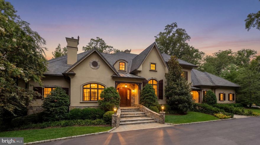 Tucked behind automatic wrought-iron gates along a picturesque, verdant stretch of country lane, 9250 Persimmon Tree Road is a spectacular French country estate that blends old-world charm and grandeur with modern amenities suited to contemporary luxury living. A winding driveway introduces park-like grounds spanning a full acre, surrounded by a lush, mature canopy of trees. The approach to the arched front door showcases intricate stonework and opens to reveal 9,850 square feet of beautifully appointed living space.

Rich wide-plank hardwood floors, extensive millwork, and a pervasive sense of craftsmanship and solidity prevail throughout. The dramatic double-height foyer features a sweeping staircase with elegant wrought-iron and wood railings. Double doors open into a private study with an intricate wall of arched built-ins, also echoed in the striking double-height living room opposite the foyer. From there, the home flows seamlessly into the embassy-sized dining room highlighted by a tray ceiling and accent lighting.

A transverse gallery connects the home’s formal spaces with more relaxed living areas on the main level. These include a great room that opens onto the covered rear deck and shares a two-sided fireplace with the more intimate family room. The chef’s kitchen is thoughtfully designed with dual islands, one with integrated seating, furniture-grade contrasting cabinetry, marble floors, and a comprehensive suite of commercial-grade Thermador appliances. Service areas are conveniently tucked just beyond and include a butler’s pantry with a wine refrigerator, a family foyer, a second powder room, and access to the oversized heated four-car garage.

A cozy vestibule introduces the main level primary suite through double doors. Occupying a dedicated wing of the home, the bedroom features a fireplace and a soaring 12-foot tray ceiling. The dressing area is flanked by two walk-in closets lined with custom built-ins. The primary bath showcases marble floors, double vanities, a jetted soaking tub set against tranquil wooded views, a separate water closet, and an oversized walk-in shower designed as a steam room with dual steam generators and a sealed glass enclosure.

Four additional bedrooms are located on the upper level, each offering generous storage and a private en-suite bath. One of these suites is particularly versatile and well suited as a hobby room, home office, or private retreat. A conveniently located laundry room completes the upper level.

The walkout lower level is anchored by a large recreation room with a fully outfitted wet bar. Additional entertaining spaces include an adjacent billiards room, a wine room with built-in storage, and a professional home cinema. A fitness center, two bedrooms, and a full bath with direct pool access complete the level, creating an ideal environment for entertaining and extended stays.

The outdoor living spaces are a true highlight. The lower level opens to a covered terrace leading to an expansive flagstone patio surrounding the 45-foot lap pool, complete with a spa and automatic cover. Elegant hardscaping and curated landscaping create a serene and private setting.

At the far end of the lawn, a pavilion with ceiling-mounted heaters and a fireplace allows for year-round al fresco entertaining. Adjacent to the pavilion, the hardscape extends to a large stone firepit area, creating an inviting gathering space for evenings outdoors. After sunset, the grounds are illuminated by landscape lighting, and the property sits on a level, fully usable lot with underground irrigation.

Despite its tranquil setting, the estate enjoys a prime close-in location less than a mile from Potomac Village and near major commuter routes into the city, while belonging to the highly sought-after Winston Churchill High School cluster.

Immaculately maintained and defined by an elevated level of style and craftsmanship, 9250 Persimmon Tree Road represents estate-style luxury at its finest.