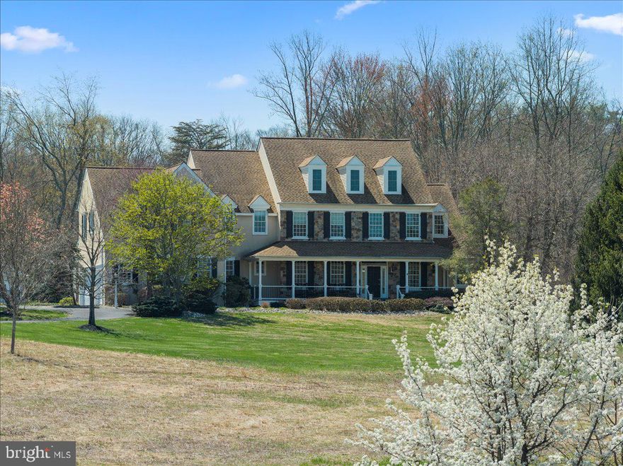 Welcome to this stunning Bucks County estate where timeless craftsmanship, natural beauty, and refined living converge on 5 breathtaking acres within the coveted Council Rock North school district. Set gracefully beyond a meadow of pollinators and framed by a wraparound porch, this custom masterpiece invites you to savor sunrise mornings overlooking your private landscape. Every detail has been thoughtfully curated to create a residence that is grand in scale and intimate in comfort. A soaring 2-story foyer with dramatic turned staircase, rich wainscoting, thick crown molding, arched hallways with keystones, and transom windows introduces interiors bathed in natural light. Hardwood floors flow throughout the main level, leading into a sophisticated formal living room adorned with a coffered ceiling and gas fireplace. French doors open to a serene deck echoing gentle sounds of the nearby Neshaminy Creek. The chef’s kitchen is nothing short of extraordinary, designed for culinary artistry and elegant entertaining. Granite counters, knotted pine cabinetry, Sub-Zero refrigerator, 48” gas range, pot filler, double ovens, wine refrigerator, stand mixer appliance lift, and an expansive island with butcher block carving station anchor the space. A tray ceiling breakfast room wrapped in windows provides the perfect setting for quiet gatherings. The great room impresses with its cathedral ceiling, wood-burning fireplace, and architectural trim, creating a warm yet dramatic space for connection. This wing of the home is enhanced by a walk-in pantry, coffee station or dry bar, well-appointed laundry room with folding station, powder room, and direct access to the oversized three-car garage. On the opposite wing, formal entertaining shines in the elegant dining room. An executive office with custom cherry cabinetry, an additional powder room, and walk-in closet contributes to sophistication and functionality. Upstairs, hardwood floors continue through the hallways accessed by both formal and casual staircases. The primary suite is a haven, featuring a tray ceiling, extended sitting area, vanity station, and dual walk-in closets. The spa-inspired bathroom boasts separate vanities, a soaking tub, private water closet, and an expansive shower with dual rain heads, all overlooking panoramic views of the estate grounds. A princess suite enjoys a spacious closet and its own private bath, while two additional bedrooms with walk-in closets share a thoughtfully designed Jack-and-Jill bath with separate private shower and toilet. A thoughtful loft area creates a designated homework zone or additional home office. The third-floor retreat is comprised of a wooden ceiling, dormer windows, generous closet space, and newer carpeting, offering versatility as a fifth bedroom, studio, or lounge. The walk-out daylight lower level is designed for unforgettable entertaining, with gas fireplace, recreation area for pool table and games, full bar with sink and refrigeration, and a spacious fitness room adjacent to a fourth full bath. The unfinished section features abundant storage. Outdoor living rivals a private resort. The saltwater gunite pool creates a focal point for summer gatherings, with waterfall and spacious integrated hot tub for up to twelve friends. A charming pool house includes a powder room, storage, and space for a future tiki bar or cabana lounge. At the rear of the property, a substantial pole barn with oversized doors, two deep garage bays, workshop space, and second-floor storage provides extraordinary utility for hobbyists or collectors. Newer roof (2017), HVAC system (2018), water heater (2023), robust 400-amp electrical service, and a partial house generator, ensuring peace of mind for years to come. Adjacent to preserved land, this extraordinary estate represents a rare opportunity to own a private sanctuary where luxury, land, and lifestyle align. Experience the estate that redefines elevated Bucks County living.