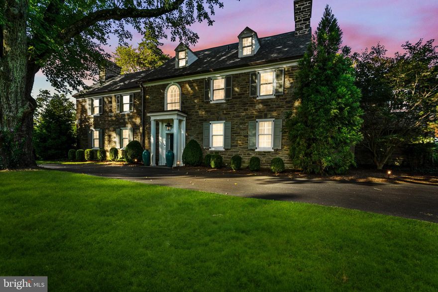 Classic and timeless, this Northside Haverford stone residence is gracefully sited on close to one acre of manicured grounds, where gardens, a shimmering pool, and tree-lined perimeters create a park-like setting. Approached by a circular drive that underscores its stately presence, the home reveals itself gradually balanced proportions, refined details, and interiors that have been thoughtfully modernized while honoring the architecture’s original spirit.

A step-up center foyer introduces a striking axial view: through the dining room’s sweeping bay window with its intricate glass roof, the eye is drawn directly to the gardens and pool beyond. To the left, the formal living room offers an atmosphere of understated elegance. An oversized fireplace, flanked by tall south-facing windows, anchors the space, while richly stained hardwood floors, continuous throughout the main level, add warmth and cohesion. Another bay window provides symmetry when viewed from the rear, and French doors open to a covered stone terrace—an invitation to flow seamlessly between interior and exterior living.

The kitchen is a study in contrast and comfort. Crisp white cabinetry plays against polished black stone countertops, while a substantial wood-topped island provides both prep and gathering space. A suite of Bosch appliances—including a steam/convection oven and dual dishwashers—makes the kitchen as functional for intimate dinners as it is for entertaining on a larger scale. Sunlight streams across the under-window sink, a small but significant detail that reinforces the room’s everyday ease.

For moments of repose, the den offers a more intimate counterpoint: custom shelving, front-facing windows, and a brick fireplace create a retreat equally suited to reading or quiet evenings by the fire.

Ascending the main staircase, an arched window becomes a focal point of architectural grace, as notable from the interior as it is when viewed from the façade. The primary suite, redesigned under current ownership, reflects both scale and serenity. The primary bath is anchored by a soaking tub positioned beneath a window, a spacious seated shower, and plenty of natural light from windows and large skylight.

A newly created walk-in closet, complete with its own heating system, offers ample storage with a sense of luxury and care.
Five additional bedrooms occupy the upper level—some connected by Jack-and-Jill baths, others with en-suite or adjoining arrangements—allowing flexibility and privacy. Anticipating the future, the attic has been spray-foam insulated, fully heated, and air-conditioned, ready for expansion should additional living space be desired.

On the lower level, daylight streams into a casual gathering area with fireplace and deep window wells—an inviting setting for informal entertaining. A secondary staircase connects this space to the kitchen, leading also to a large laundry area, half bath, and generous storage.

Outdoors, the home’s placement on its deep lot is deliberate and graceful. The covered terrace and poolside stonework blur the line between garden and architecture, while a recently re-roofed garden shed adds utility. A multi-car courtyard tucked to the rear offers discreet yet ample parking, underscoring the estate’s ability to welcome both intimate and grand occasions with equal ease.

Classic and enduring, the home is one of balance and beauty—bright yet grounded, sophisticated yet welcoming—an estate of timeless appeal in one of Haverford’s most distinguished enclaves