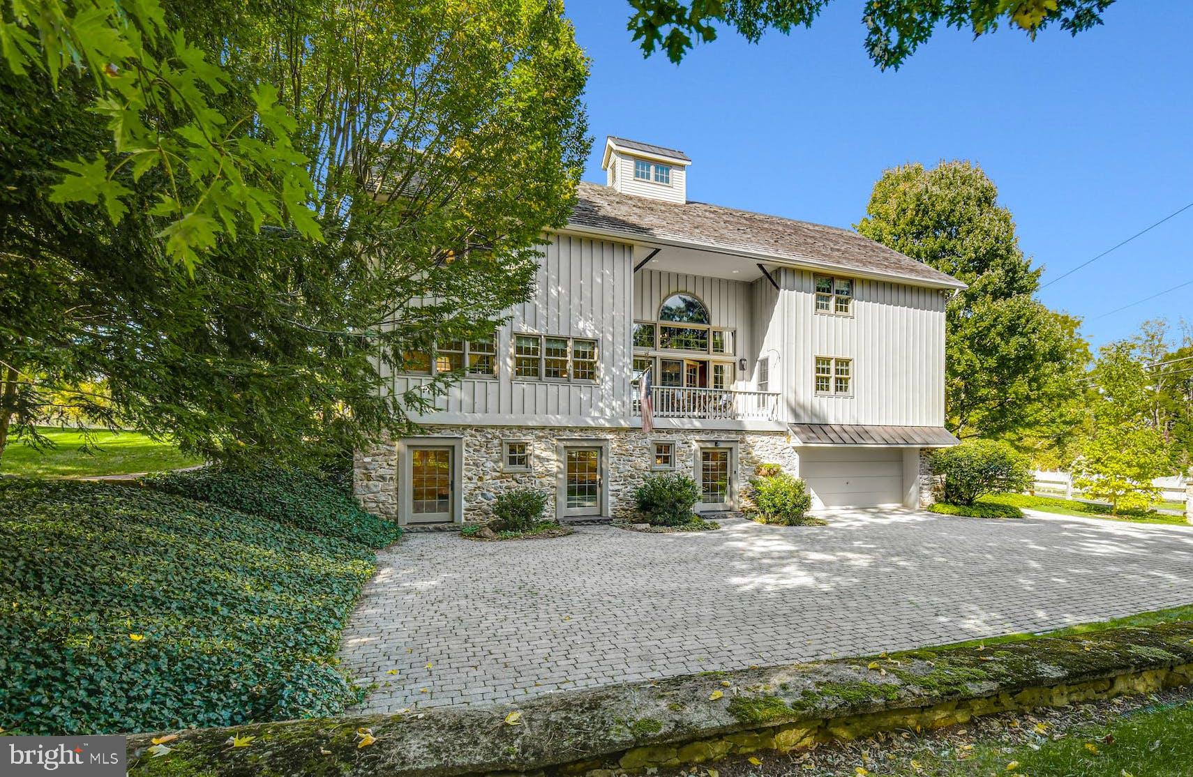 Perched above a gently winding road and surrounded by open fields and preserved lands, this circa 1850 barn was thoughtfully converted in 1988. Key interior details were left intact, including hayloft ladders, gleaming random width wood floors, and hand-hewn pegged beams. Soaring ceilings and abundant windows let in the beauty of every season.
The brick and bluestone walkway leads to a koi pond with stone waterfall, the quiet chatter of water acting as the perfect backdrop for more than 8 acres of meadows and woodland. Four-panel French doors welcome residents and guests alike into the aptly named great room, with soaring ceilings, bar closet, the first of 3 wood-burning fireplaces, and enough space for a large dining area. Adjacent, the living room and library, each with their own fireplace, offer spaces for entertaining or relaxing in the evening. The 2022 kitchen is a chef’s dream, with radiant heated floor, granite countertops and backsplash, and walk-in pantry. Laundry facilities, a half bath, and office with loft area complete this level.
Upstairs, the primary suite includes a walk-in closet and luxurious bath with console vanity, clawfoot tub, and glass shower. Three more bedrooms and two full baths all afford bucolic views of the surrounding acreage. The finished lower level with radiant heated floor serves as a multi-purpose room, with exposed stone walls and doors to the parking area. Ideally located in Upper Bucks County, with shopping, dining and commuter routes just a short drive away, Longmeadow is perfect as a full-time residence or weekend retreat.